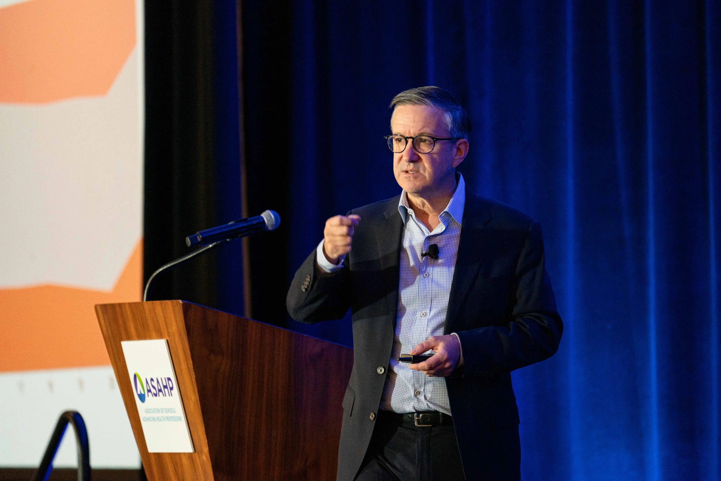 A man wearing glasses and a dark suit jacket giving a presentation at a podium with a microphone at a conference. The podium has a sign that says 'ASAH', and there is a large screen with a partly visible slide behind him. The background features dark