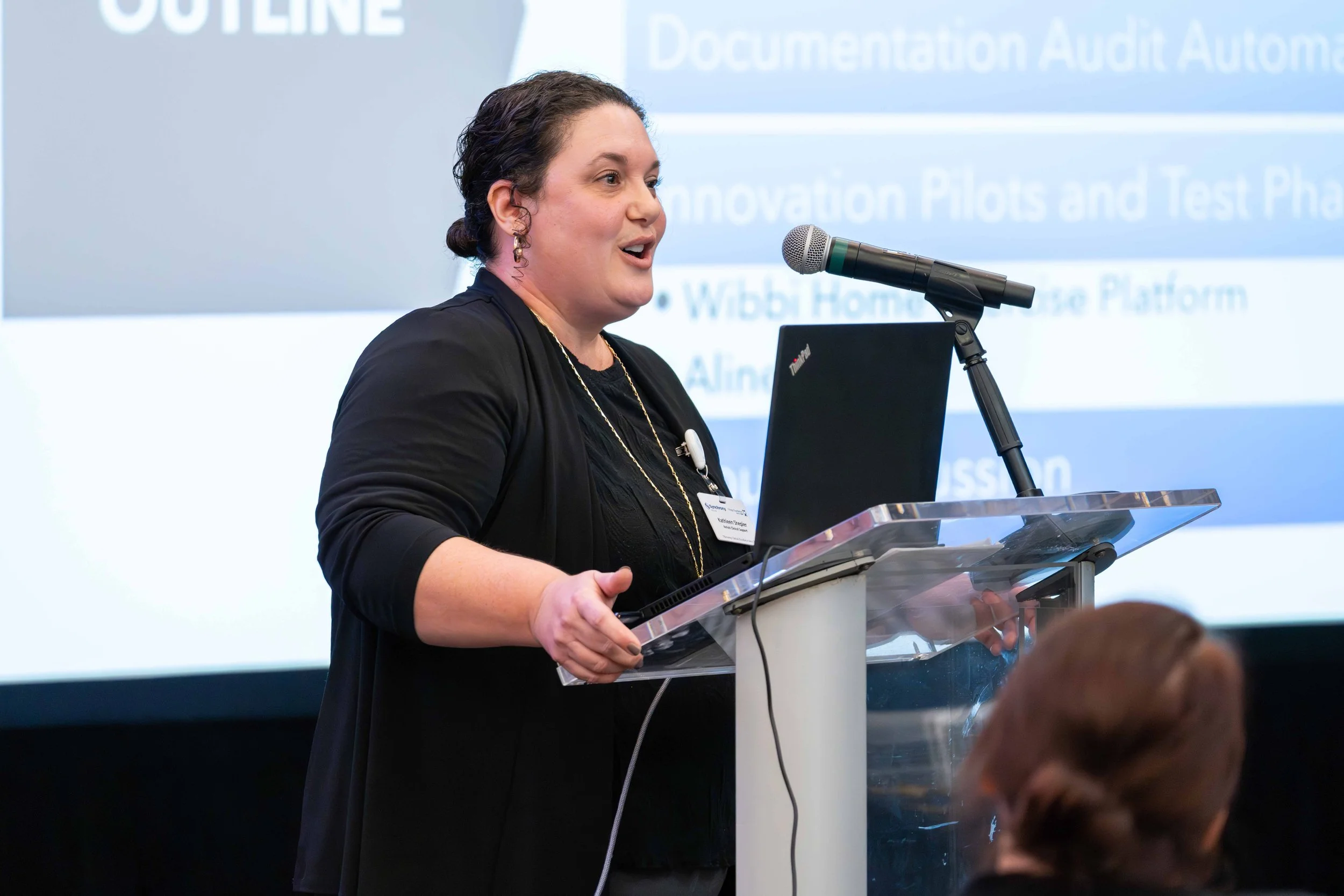 A woman speaking at a conference behind a clear podium with a laptop, with a presentation slide in the background that has text about documentation audits and innovation pilots.
