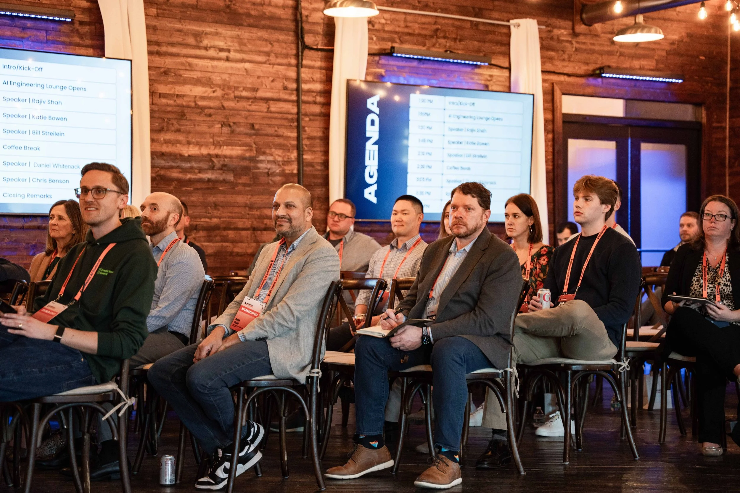 Attendees sitting in a conference room, listening to a presentation, with screens displaying agenda on the wall behind them.