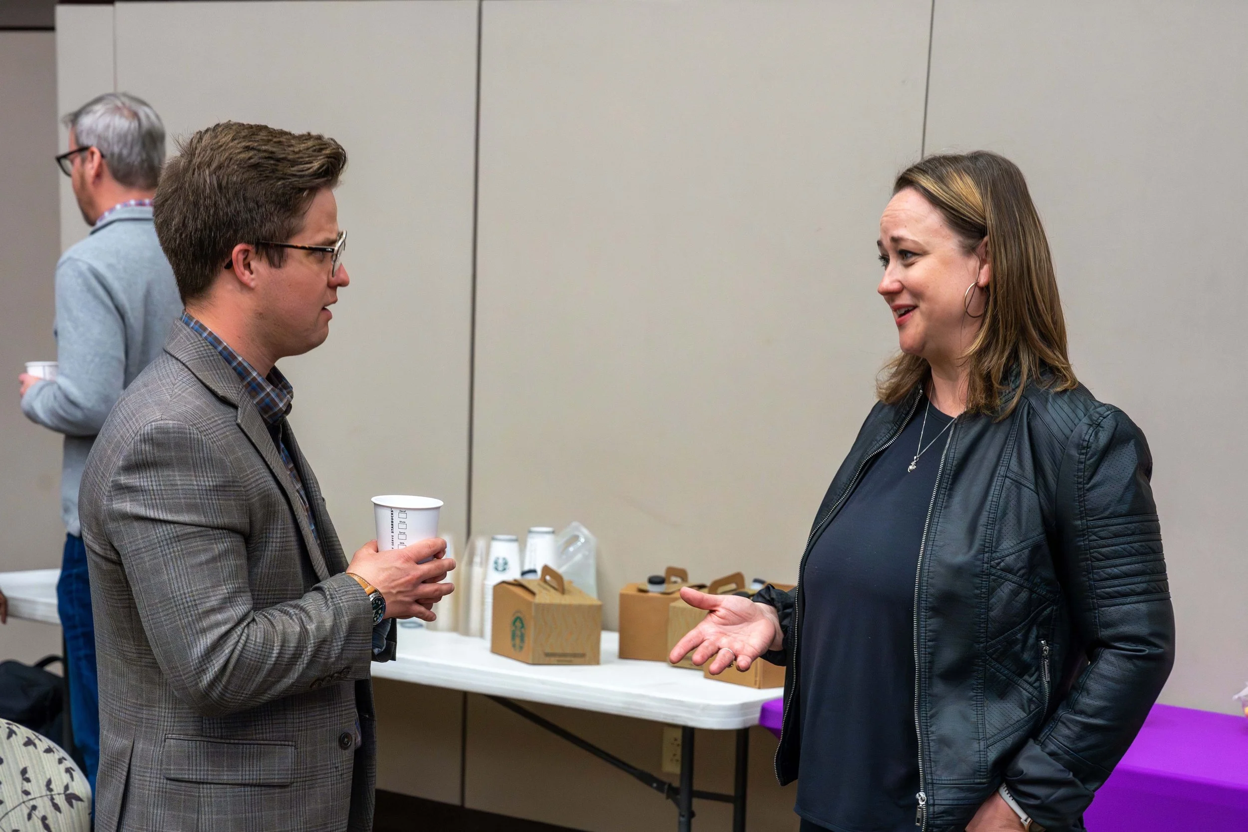 Two people are engaging in conversation at an indoor event, with a man holding a paper cup and wearing glasses and a plaid blazer, and a woman wearing a black leather jacket and earrings, expressing a point.