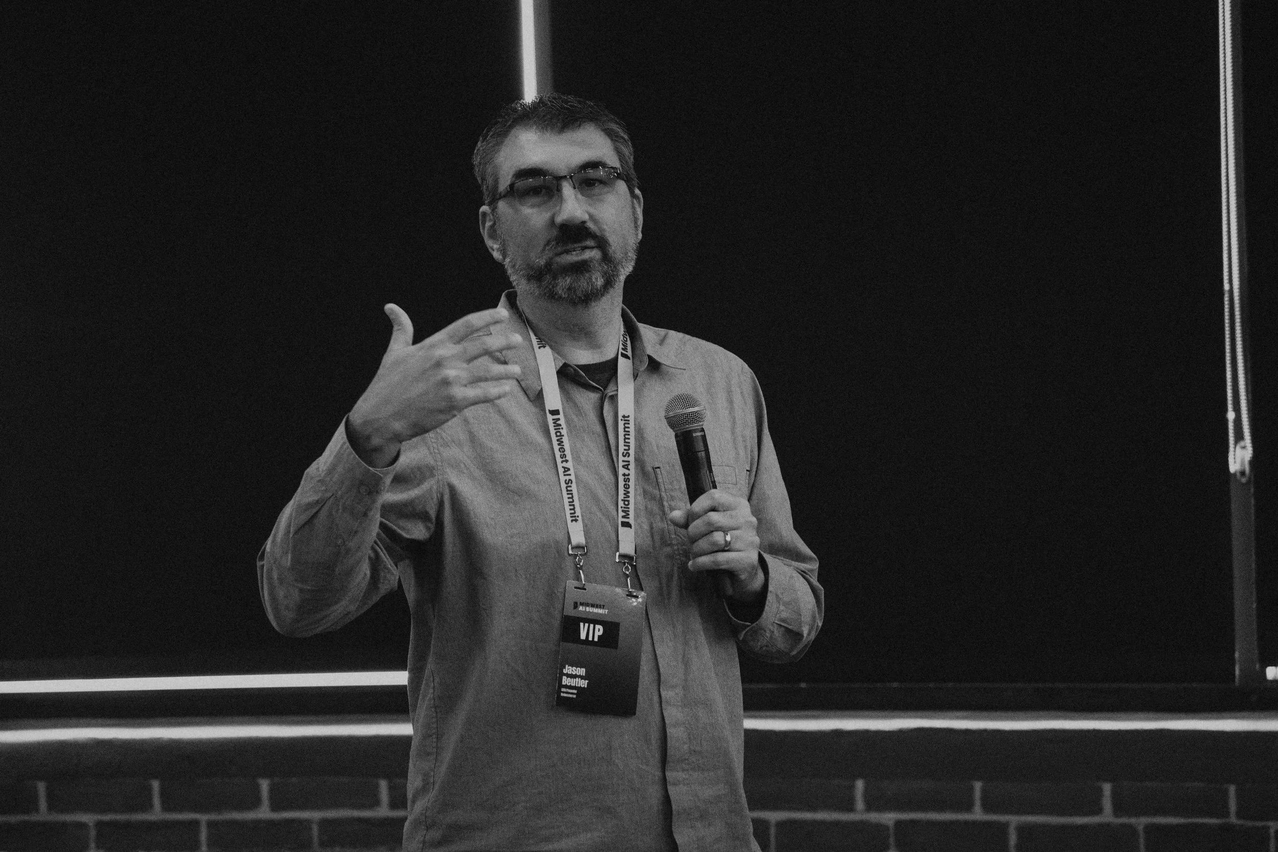 A man with glasses and a beard is speaking into a microphone, gesturing with his left hand. He is wearing a shirt and a lanyard with a VIP badge that reads "Jason Beutler" and "Midwest AI Summit." He appears to be presenting in front of a dark backgr
