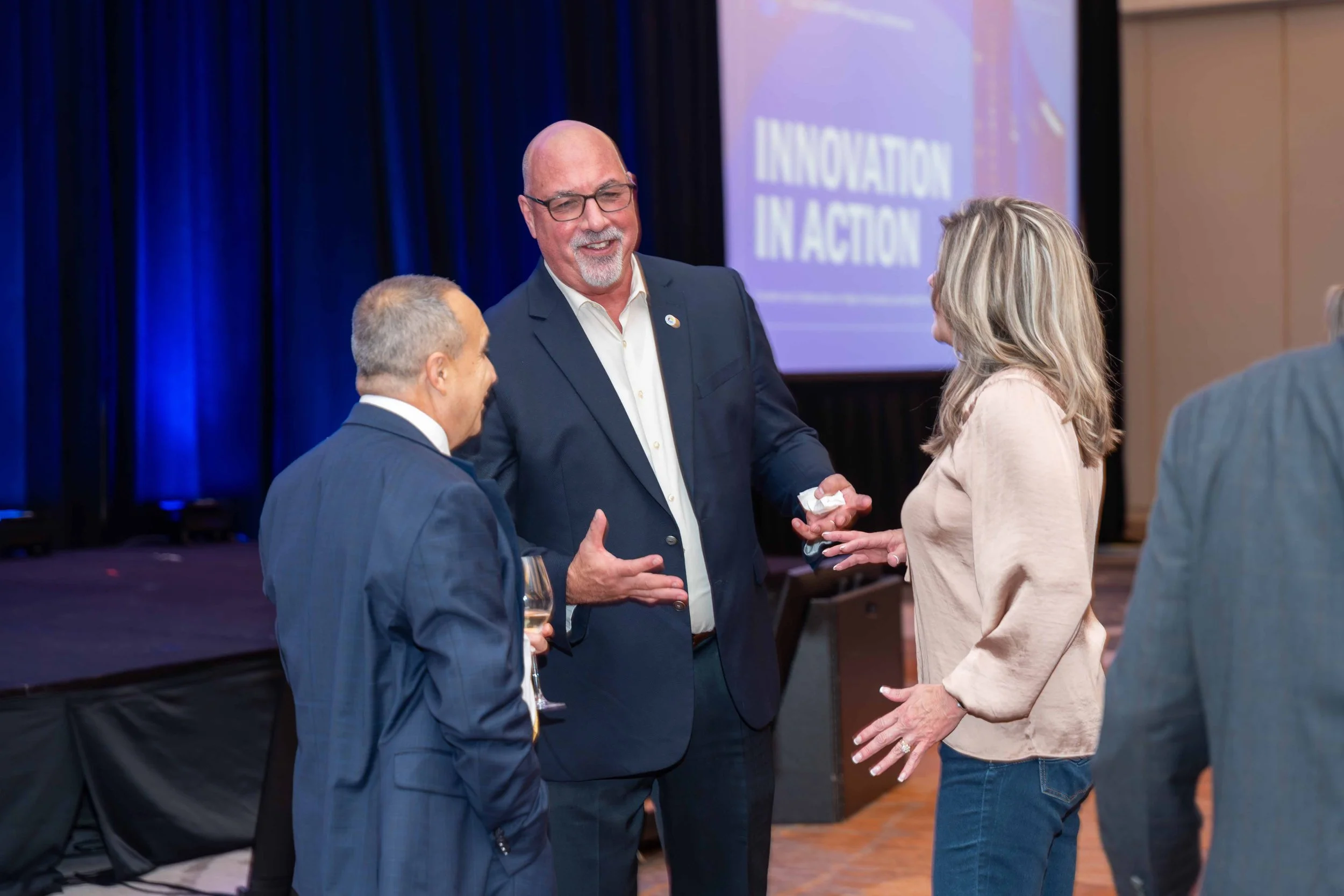 Three people engaged in conversation at a conference, with a large screen in the background displaying the words 'Innovation in Action'.