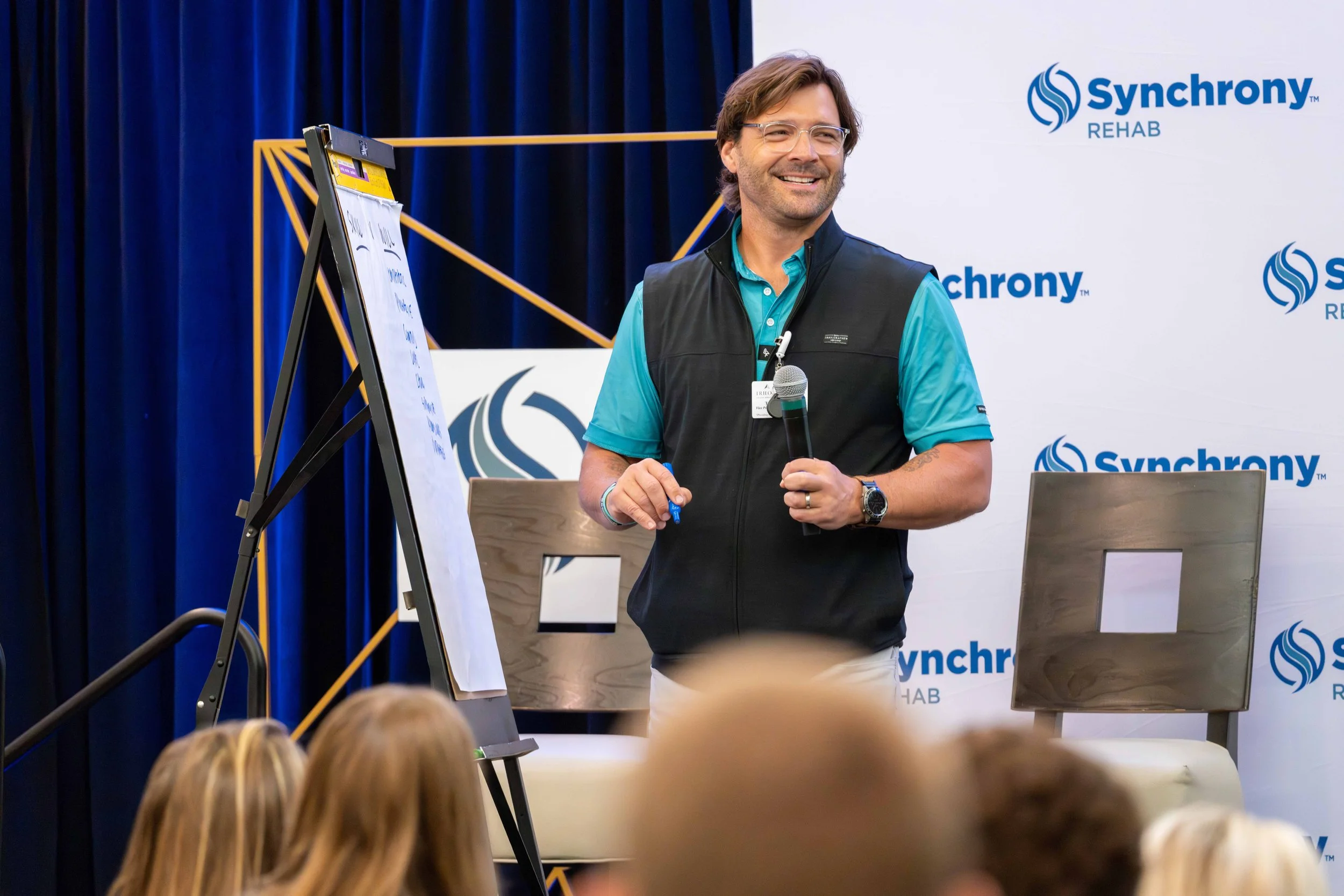 A man smiling and holding a microphone during a presentation at a conference, with a whiteboard and a backdrop displaying the 'Synchrony Rehab' logo.