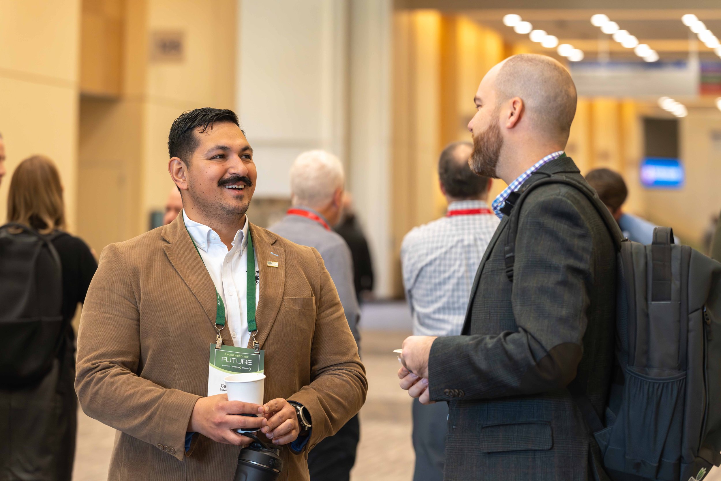 Two men are talking and smiling at a conference. One man is holding a cup and a camera, wearing a tan blazer and a white shirt. The other man has a backpack and is wearing a dark blazer, and is holding a notepad.
