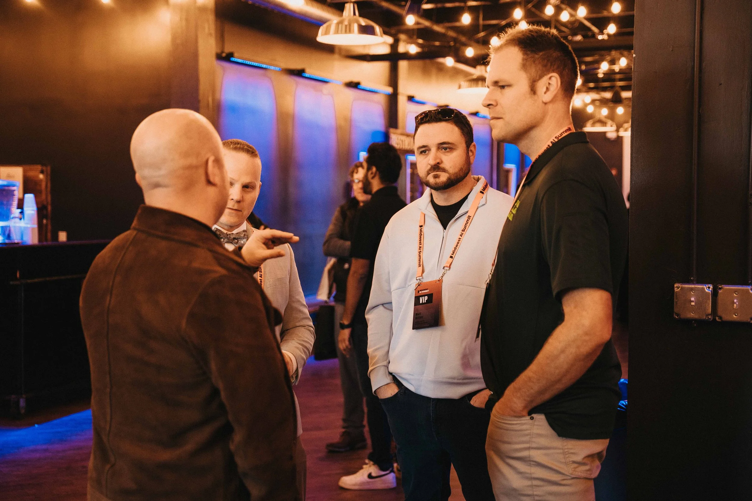 Four men engaged in conversation at an indoor event, with dim lighting and string lights overhead.