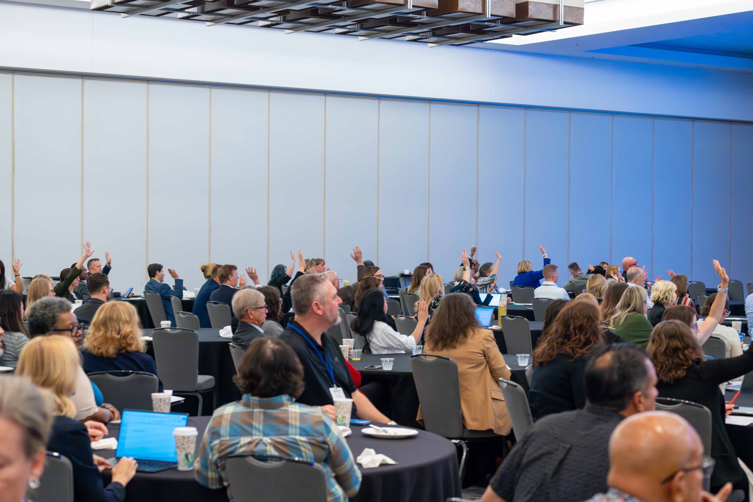 Conference room filled with people attending a meeting, many with hands raised.