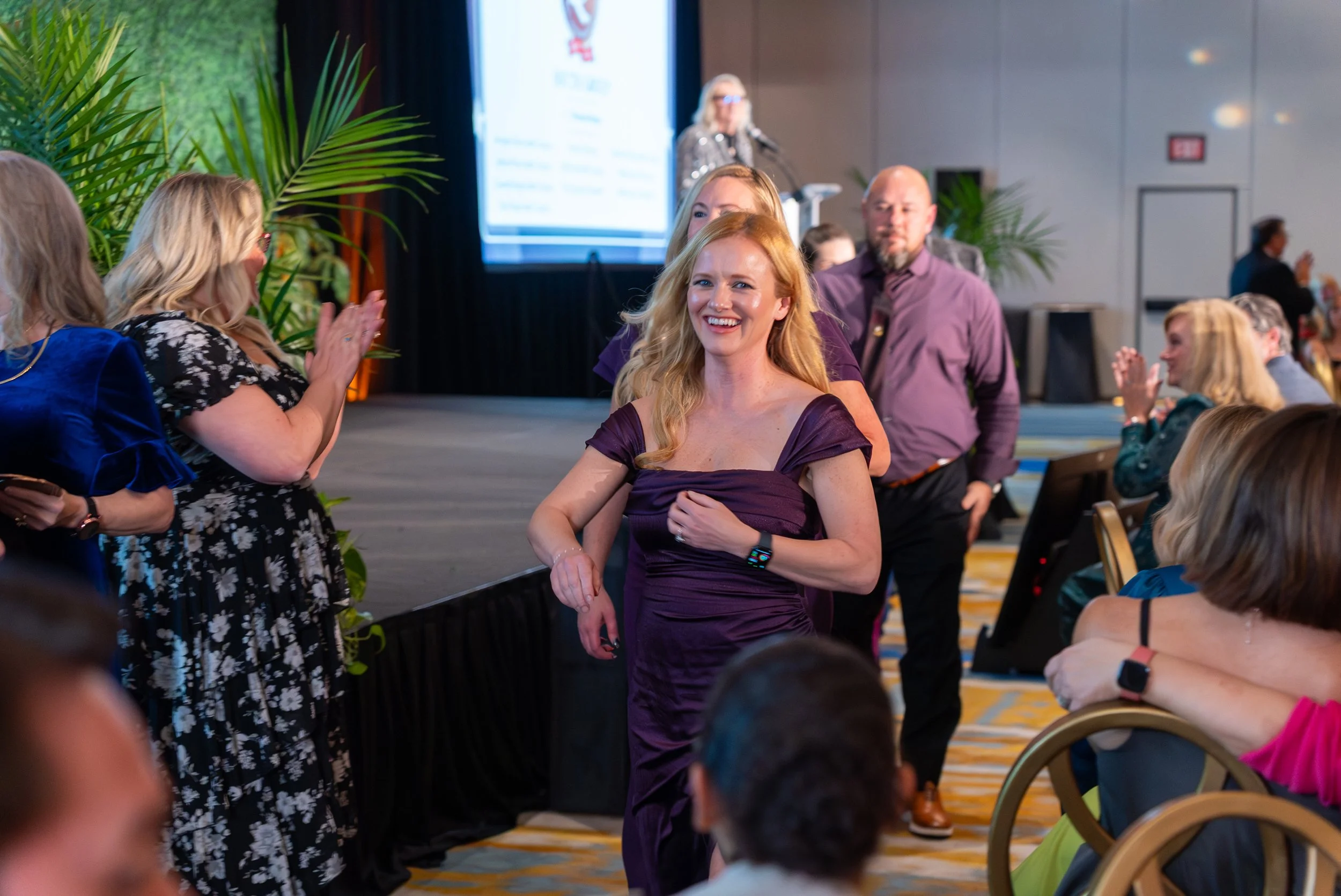 A woman in a purple dress is smiling and walking through a formal event, with people clapping and talking around her in a large conference room with a stage and projector screen in the background.