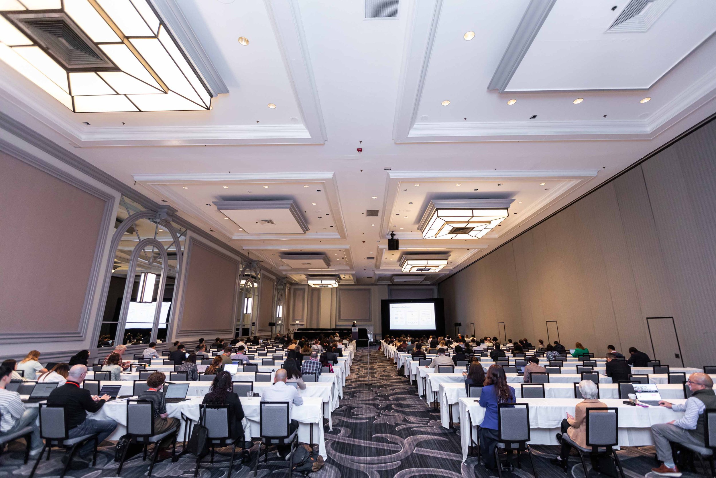 A large conference room filled with attendees sitting at long tables with white tablecloths, facing a stage with a large screen at the front. The room has high ceilings with modern light fixtures and large wall mirrors.