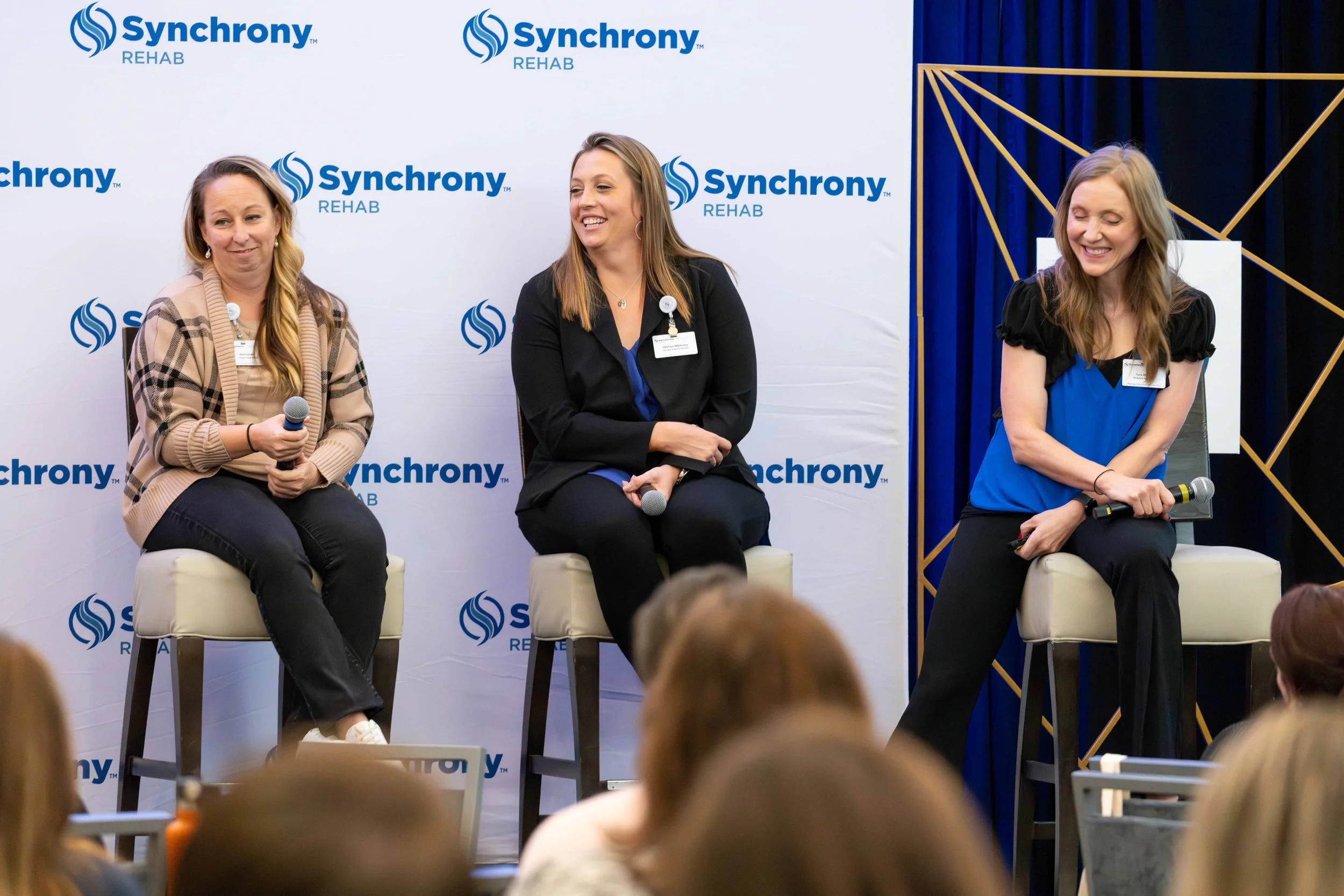 Three women sit on chairs on a stage during a panel discussion at an event, with a backdrop displaying the Synchrony Rehab logo, and an audience in the foreground.