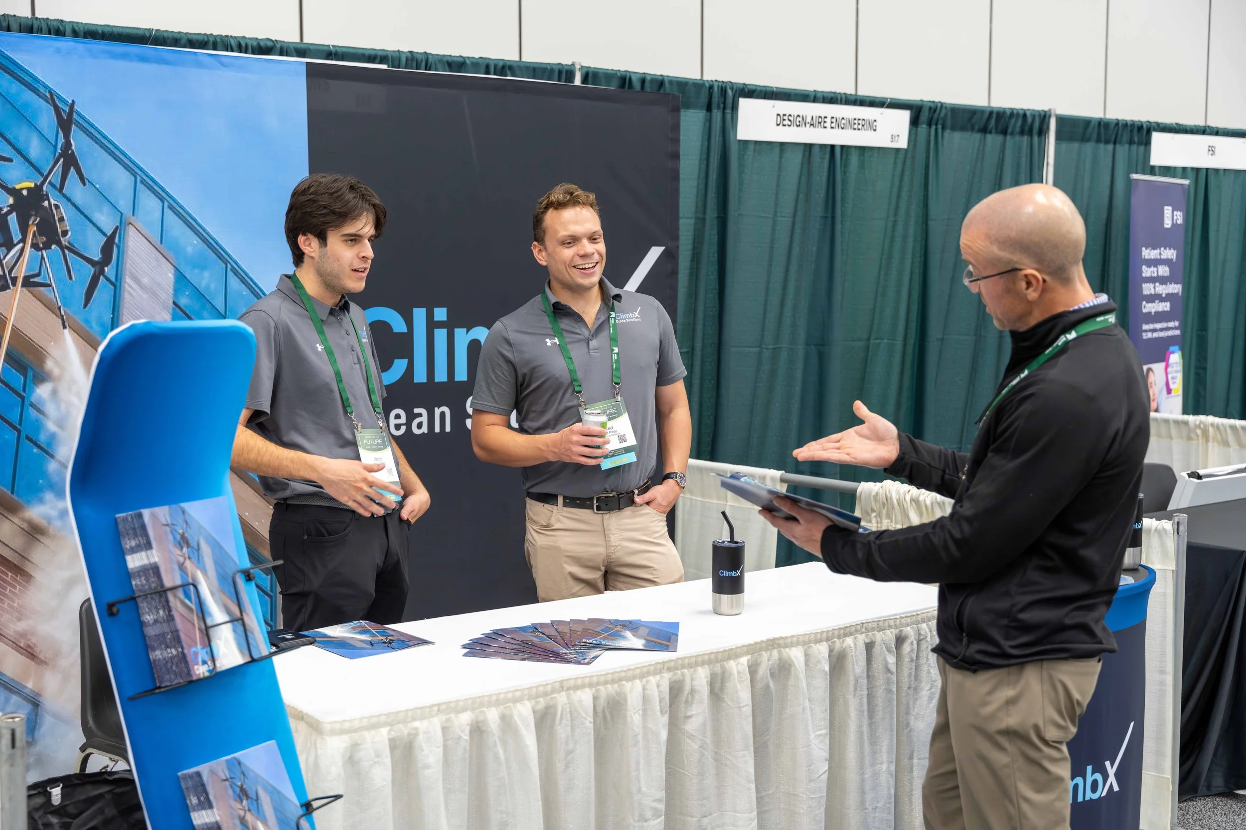 Two young men with green lanyards talking to a bald man with glasses and a black jacket at an exhibition booth for ClimbX, with brochures on the table and green curtains in the background.