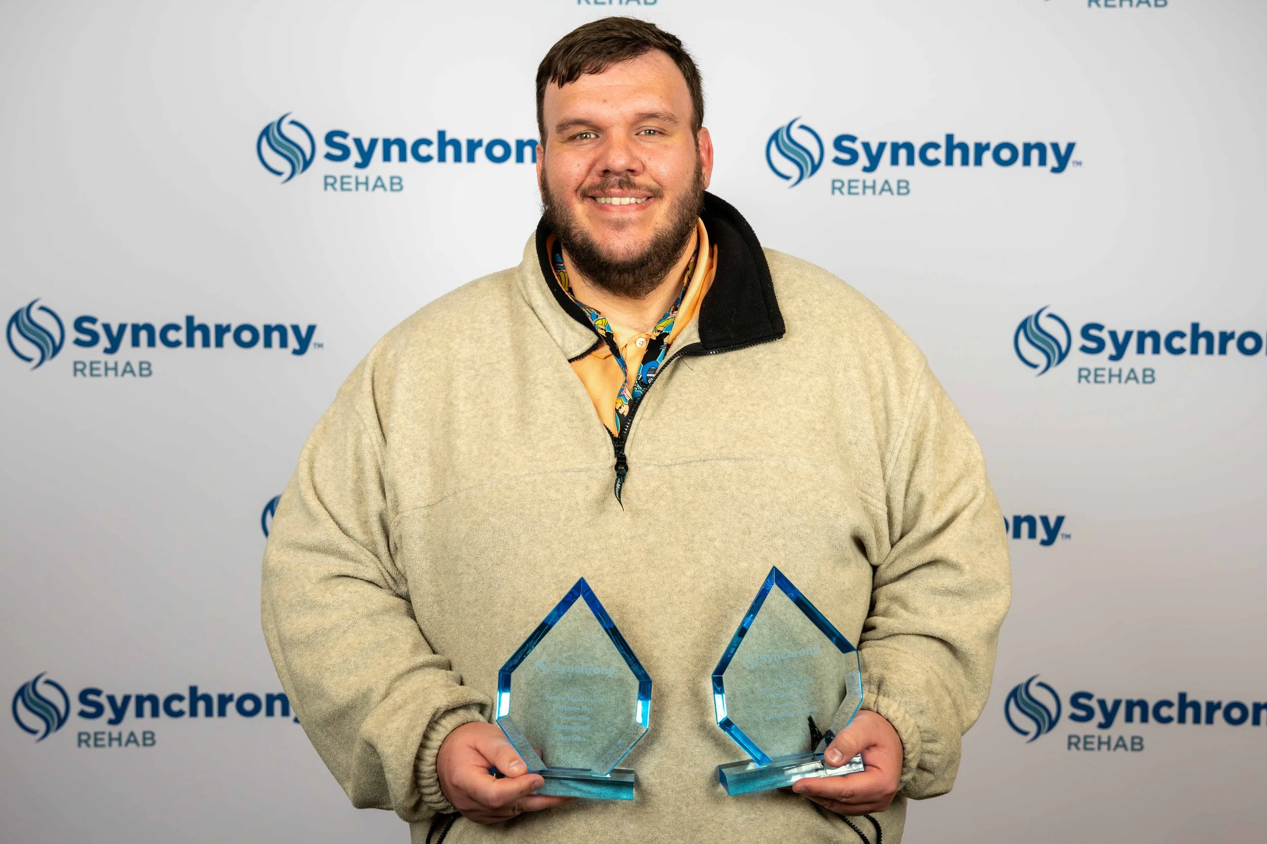 A man holding two glass award trophies standing against a backdrop with the 'Synchrony Rehab' logo repeated multiple times.