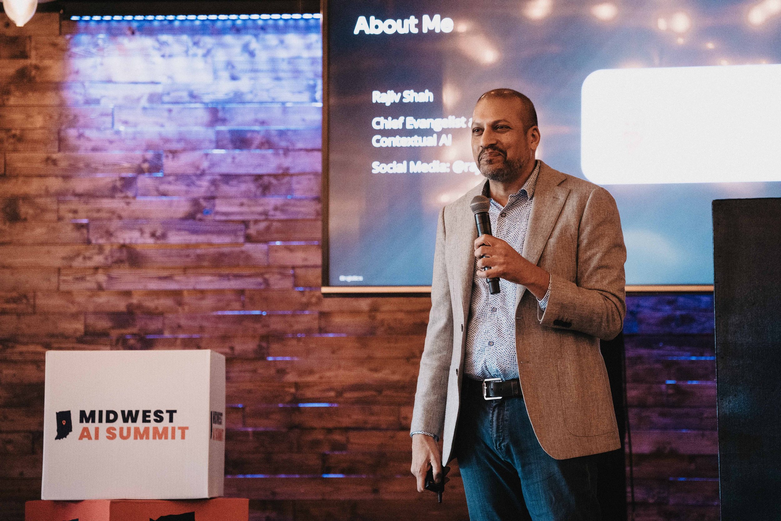 Man in beige blazer giving speech with microphone at Midwest AI Summit, standing in front of wooden wall and presentation screen.
