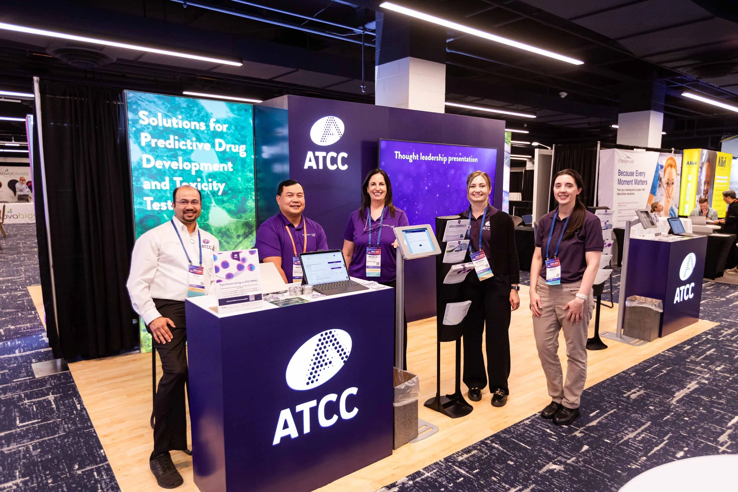 Group of five people standing behind an ATCC booth at a conference or exhibition, with digital screens displaying information about drug development and toxicity testing