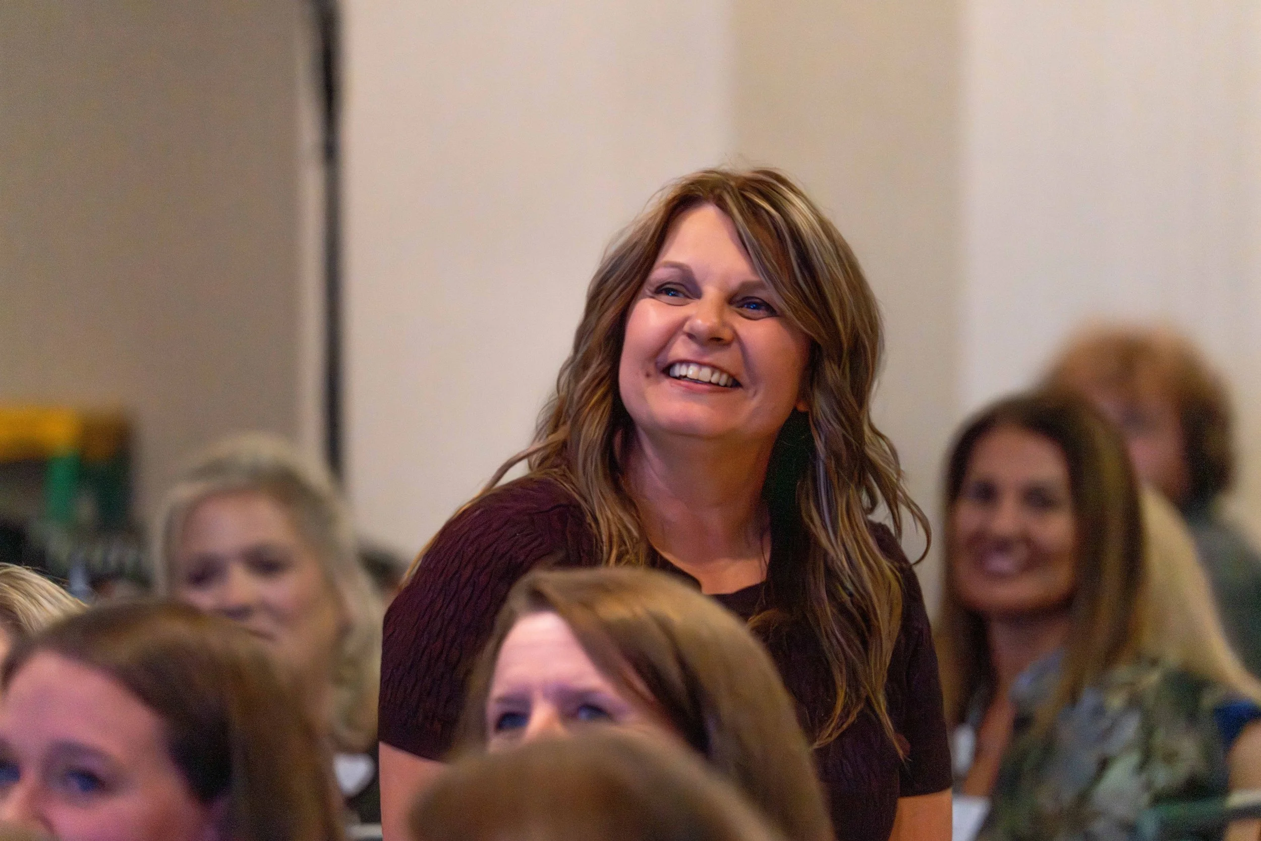 A smiling woman with long, wavy hair standing among seated people at an event.