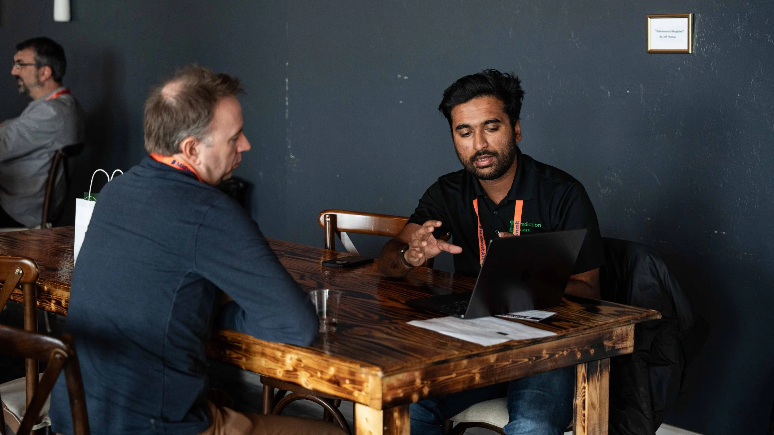 Two men sitting at a wooden table having a discussion, one is using a laptop and the other is speaking. Another person is blurred in the background.