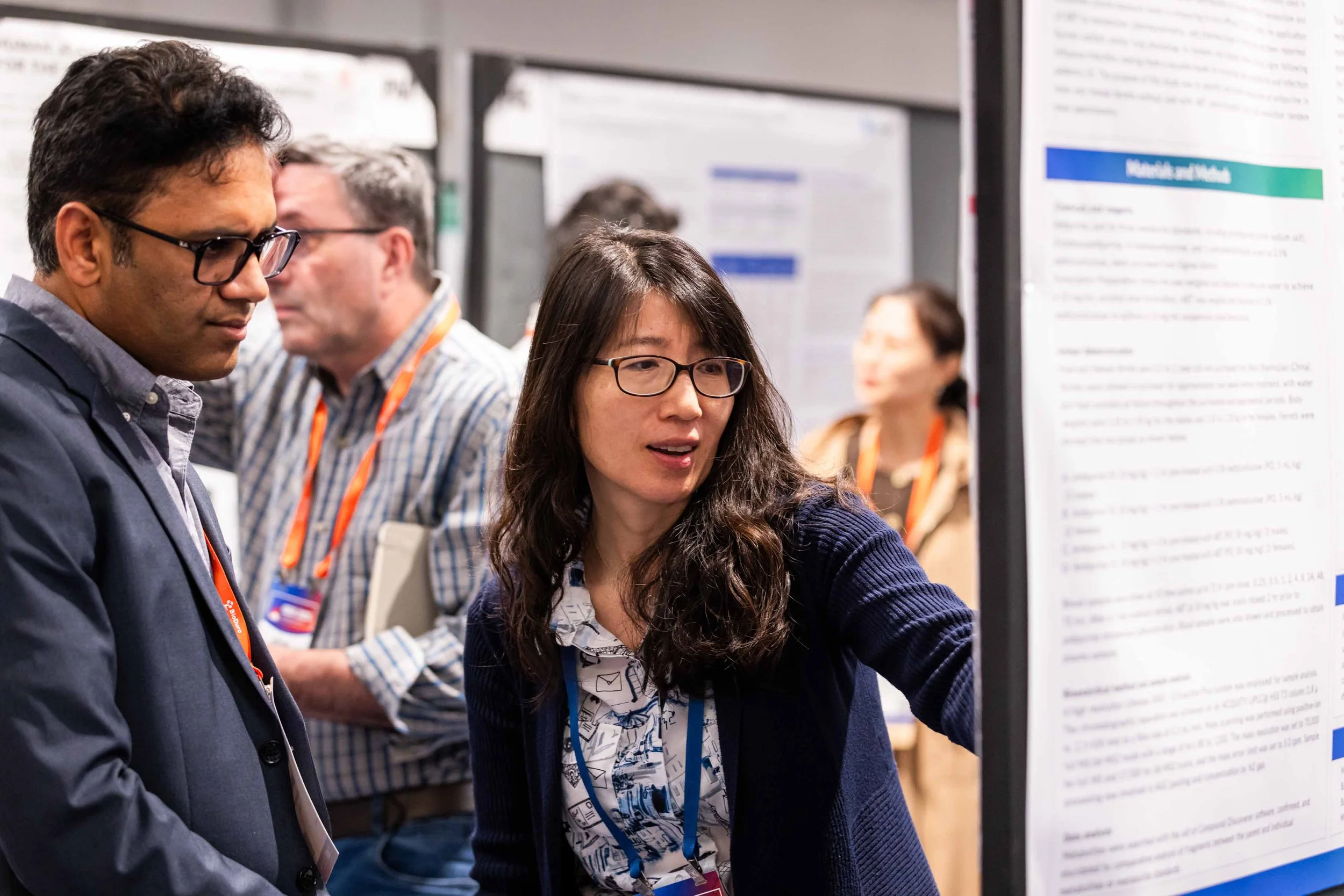 Two people, a man with glasses and a woman with glasses and wavy dark hair, are discussing a research poster at a conference. In the background, other attendees are also engaging with posters.