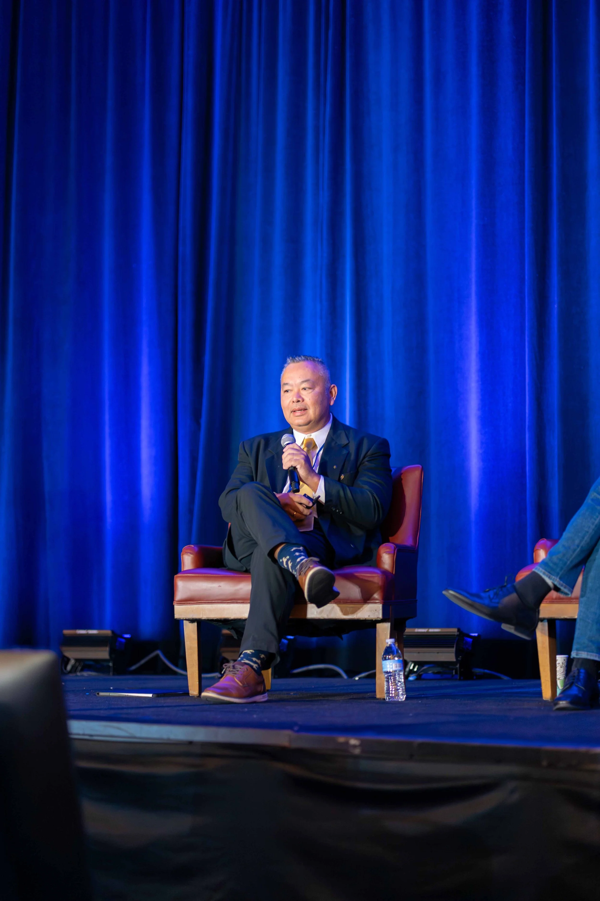Man in a blazer and dress shoes sitting on a stage chair, holding a microphone with a blue curtain backdrop.