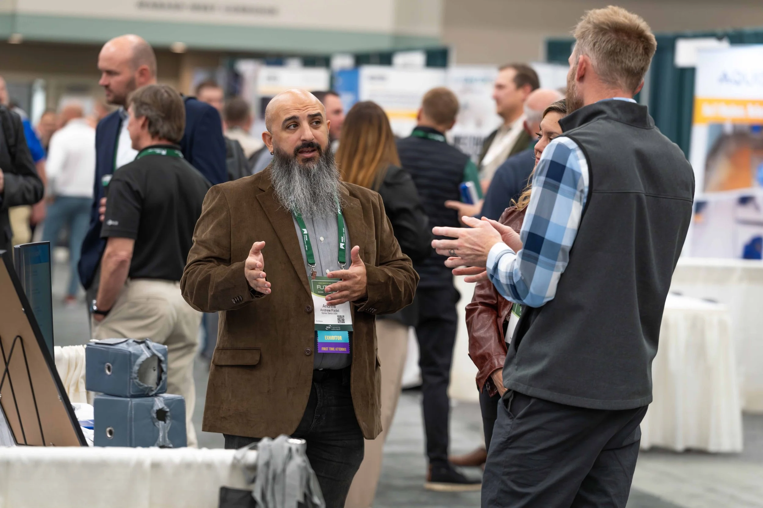 Two men having a conversation at a professional event or conference, with multiple attendees and exhibit booths in the background.