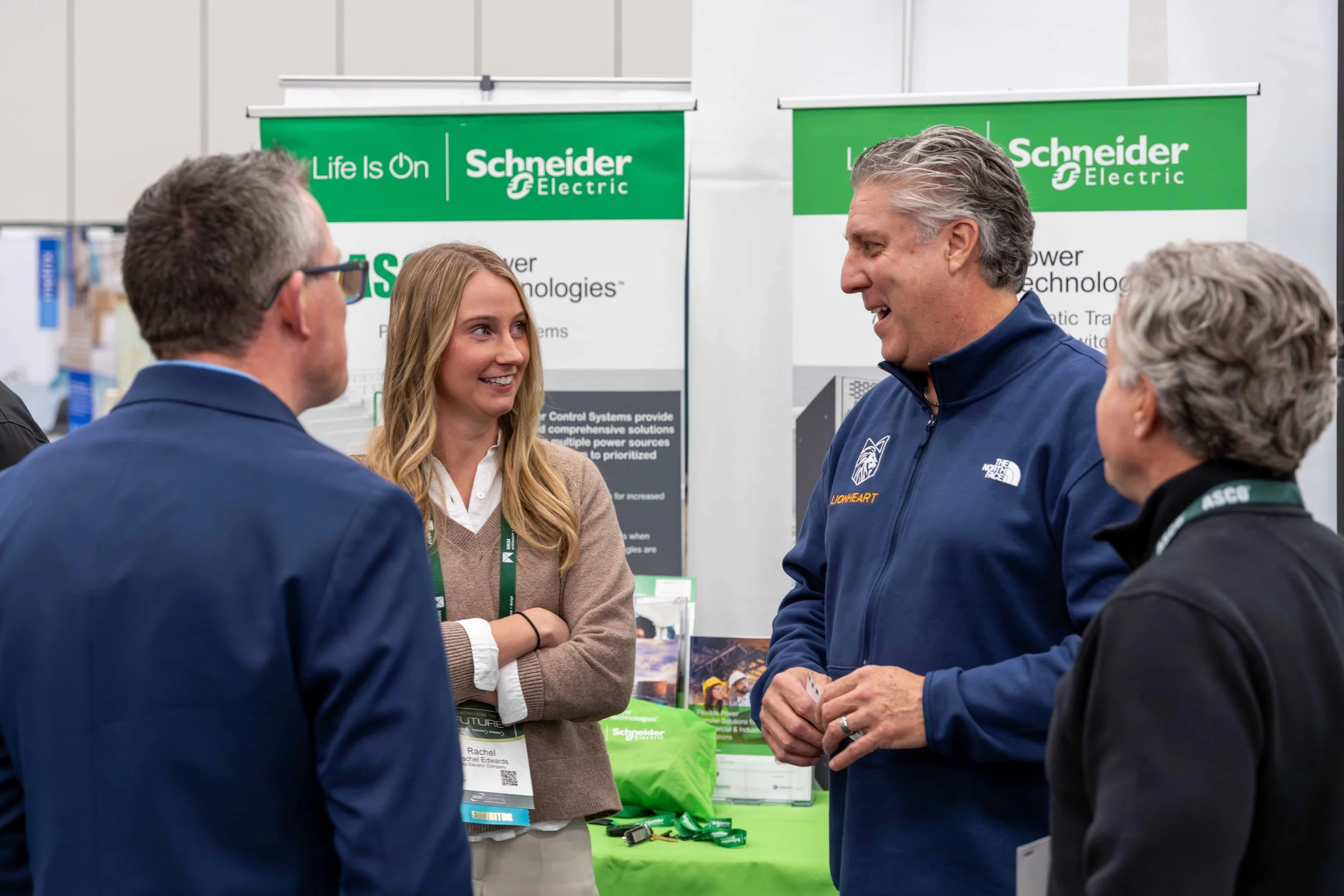 Four people engaged in conversation at a trade show booth for Schneider Electric, with banners displaying the company's logo and information in the background.