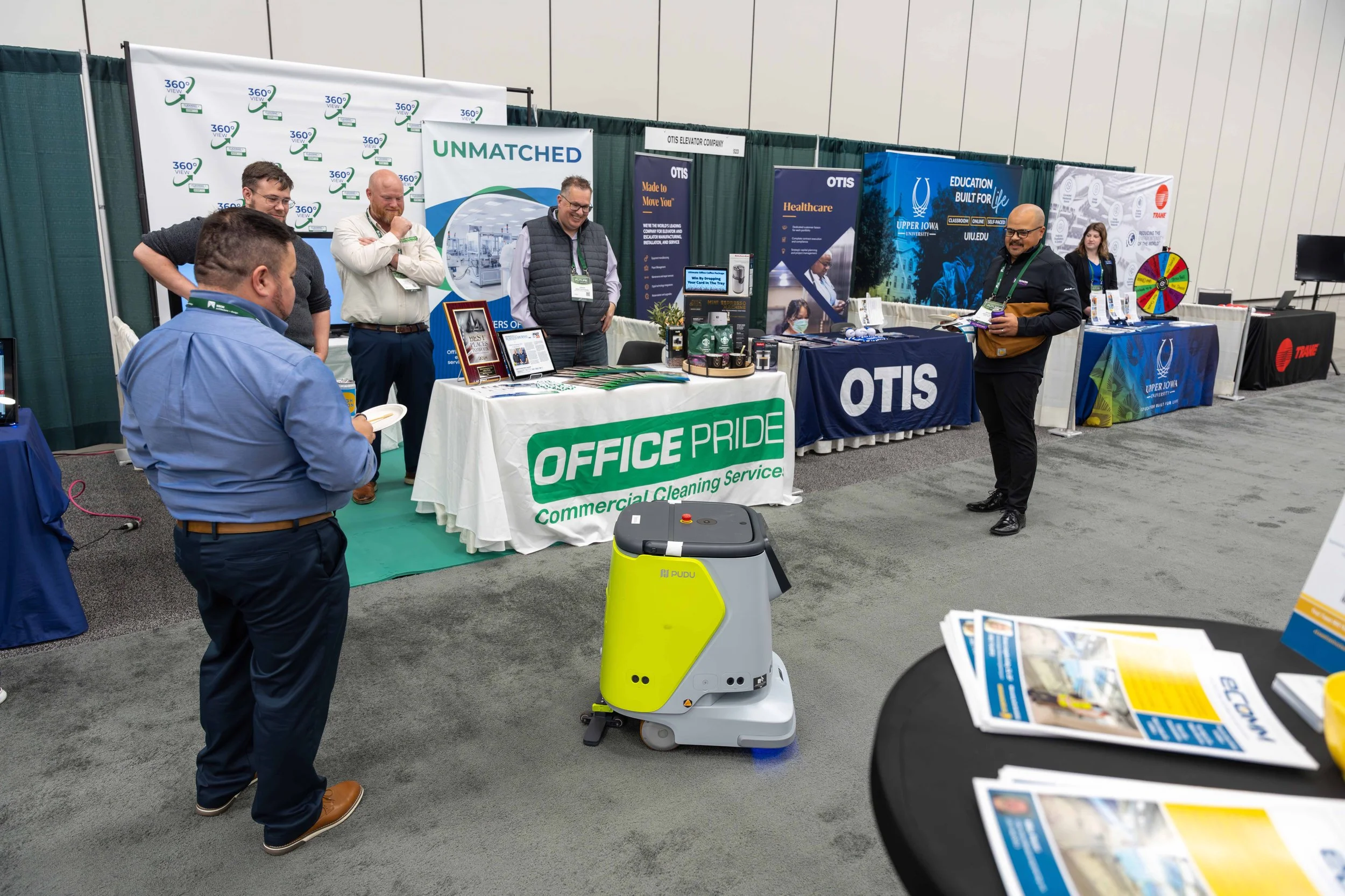 At an indoor trade show, several booths are set up with banners and promotional materials. A yellow cleaning robot is on the floor, with people standing around it, engaging and observing. Each booth displays company names and products, including Offi