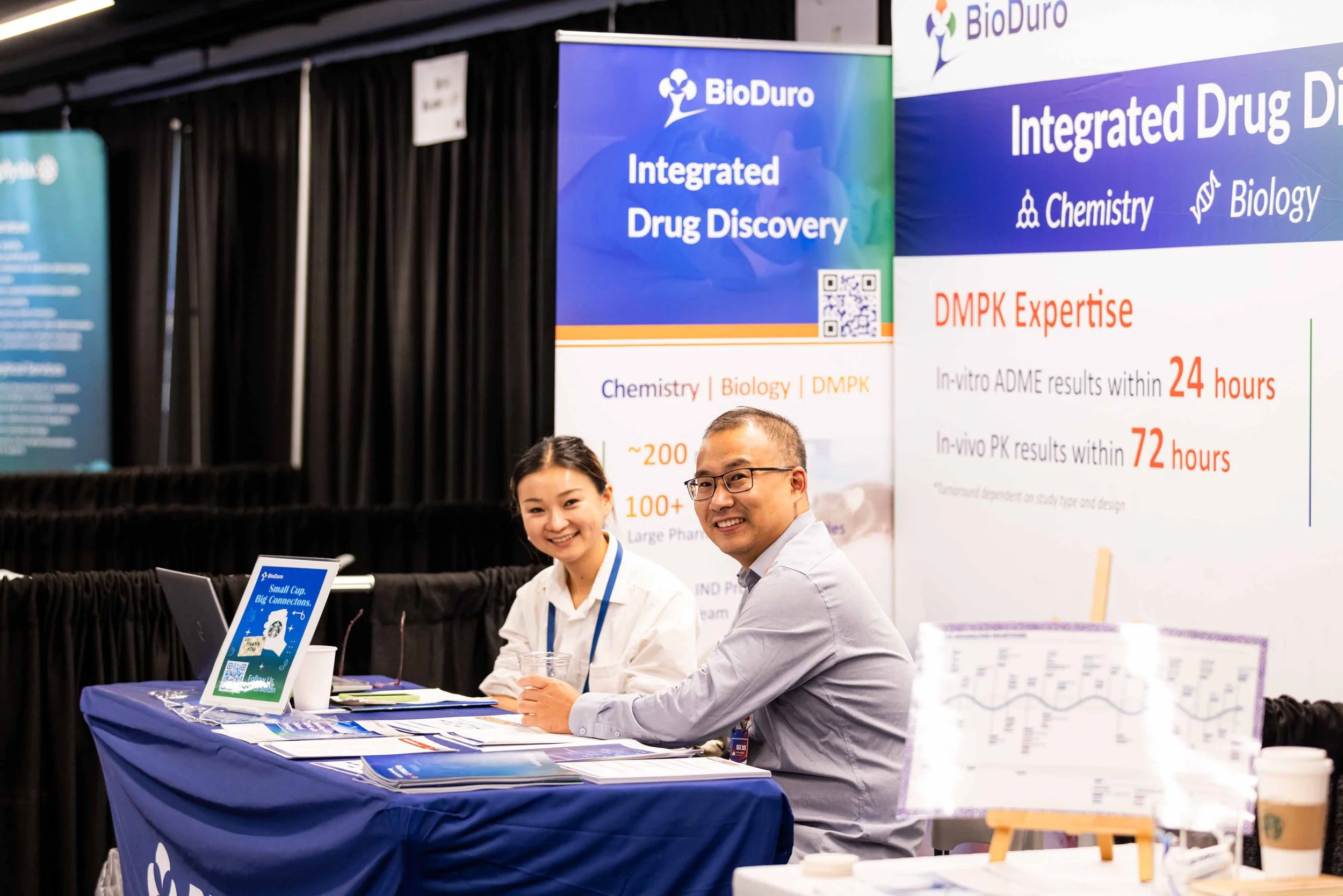 Two smiling people sitting at a table with promotional banners in the background at a conference or trade show, promoting integrated drug discovery services.
