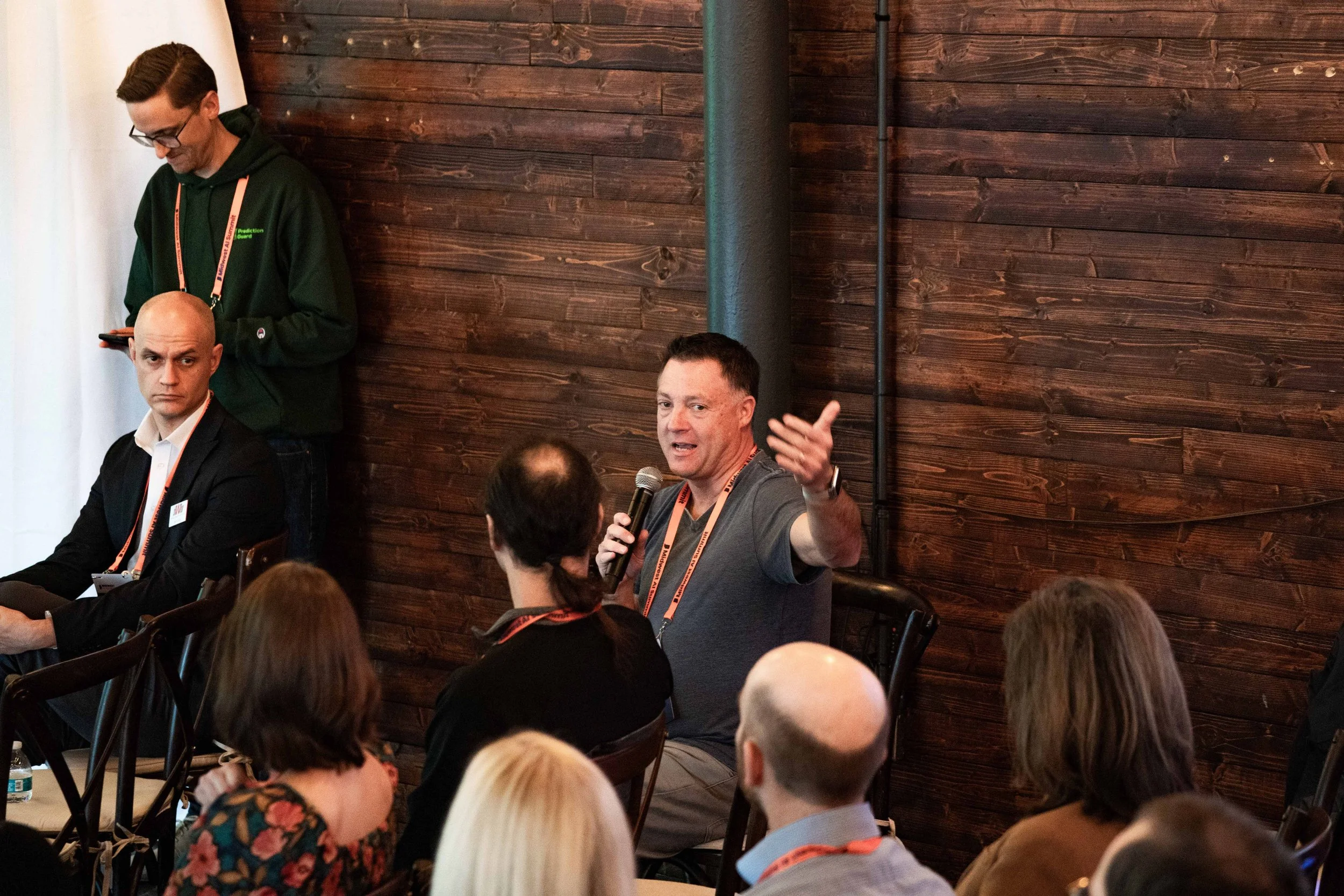A man in a gray shirt is speaking into a microphone while gesturing with his hand during a panel or conference. A seated audience listens, and there are other panelists and a person standing in the background against a wooden wall.