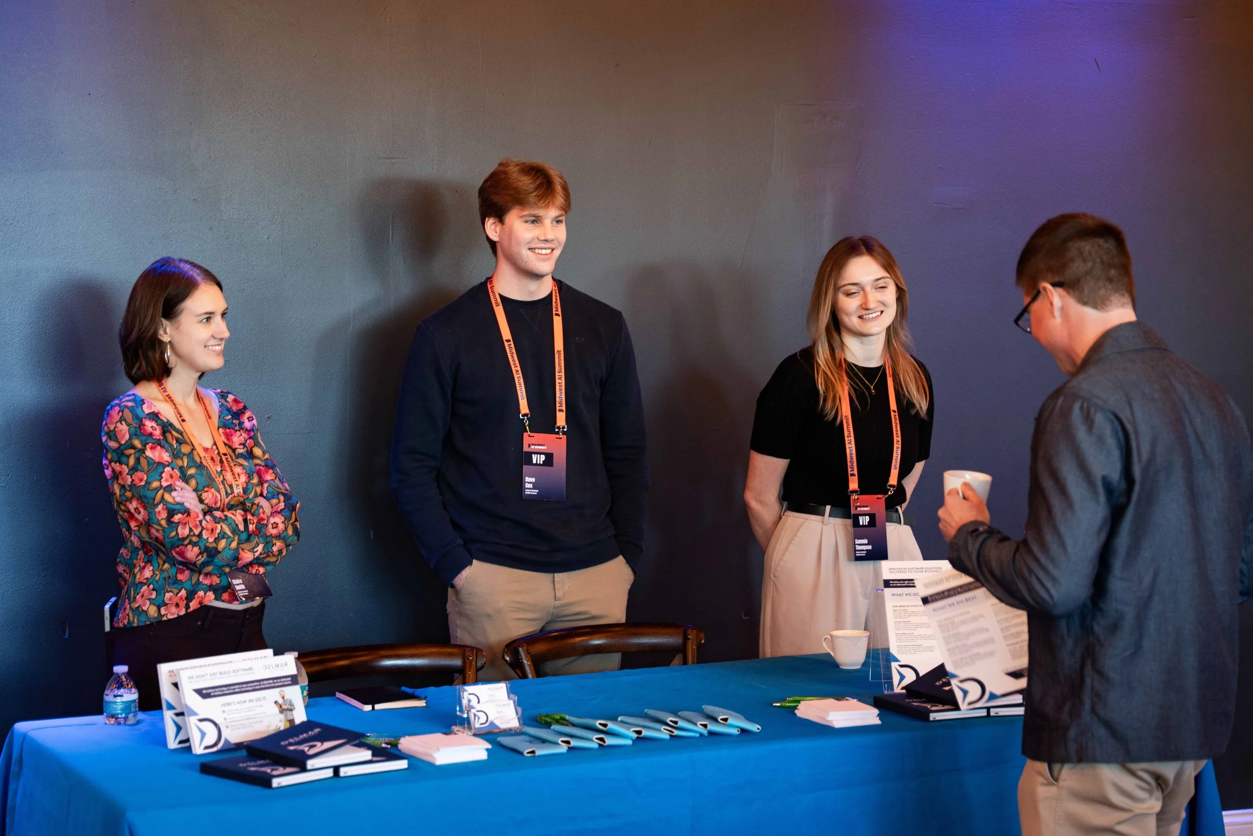 Four people stand behind an event registration table, smiling and talking to a man holding a cup. The table has pamphlets, notebooks, pens, and a water bottle. The wall behind them is plain and dark, with purple and orange lighting.