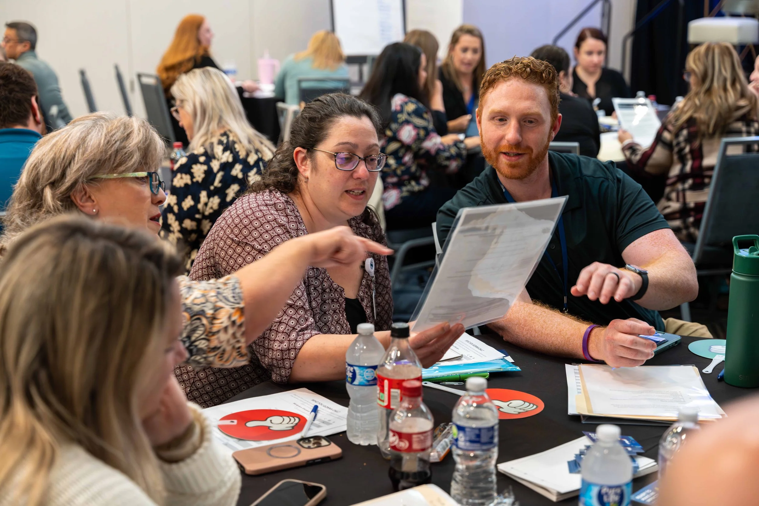 People sitting at a round table participating in a discussion or meeting, with documents and water bottles on the table.