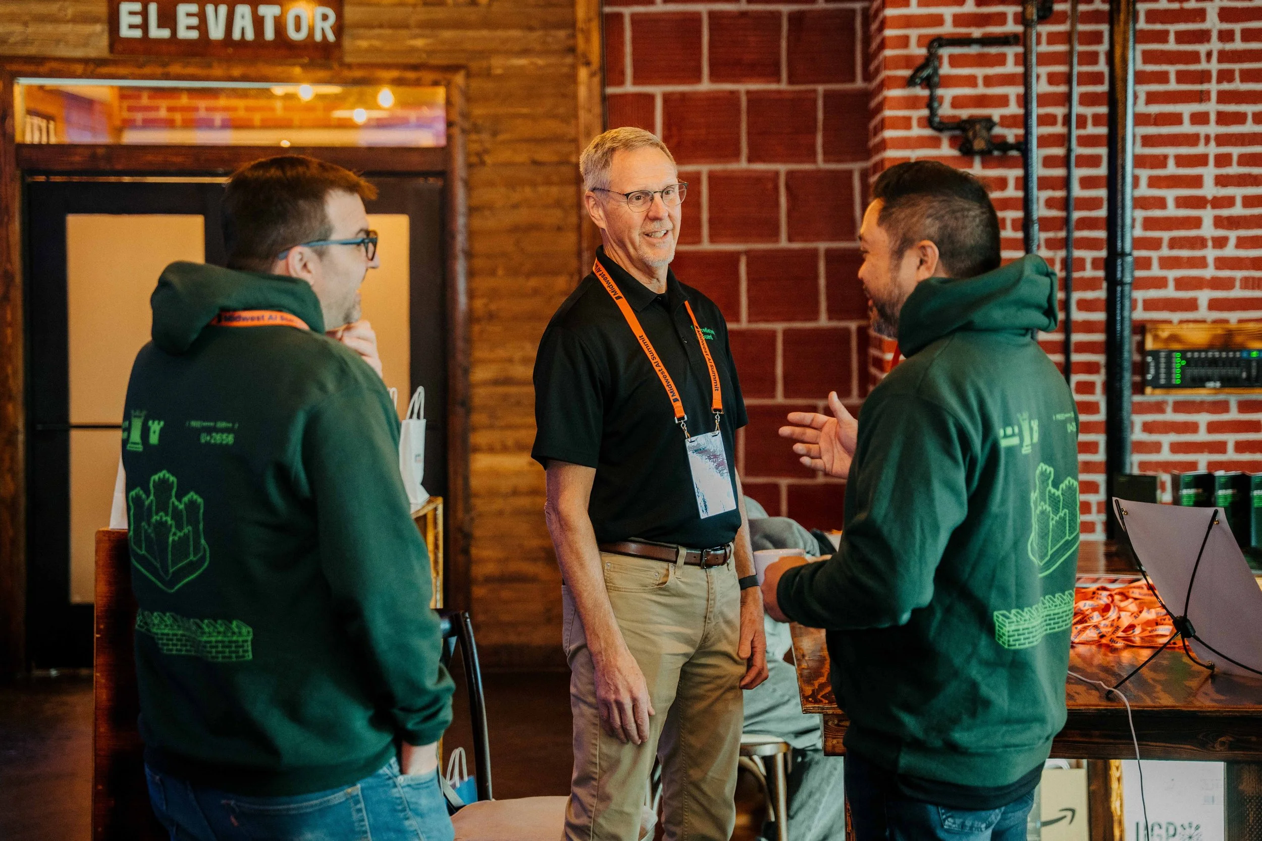 Three men in conversation at an indoor event, one in the center with glasses and a black shirt, two others wearing green hoodies, in a space with brick and wood walls, and an 'ELEVATOR' sign above.