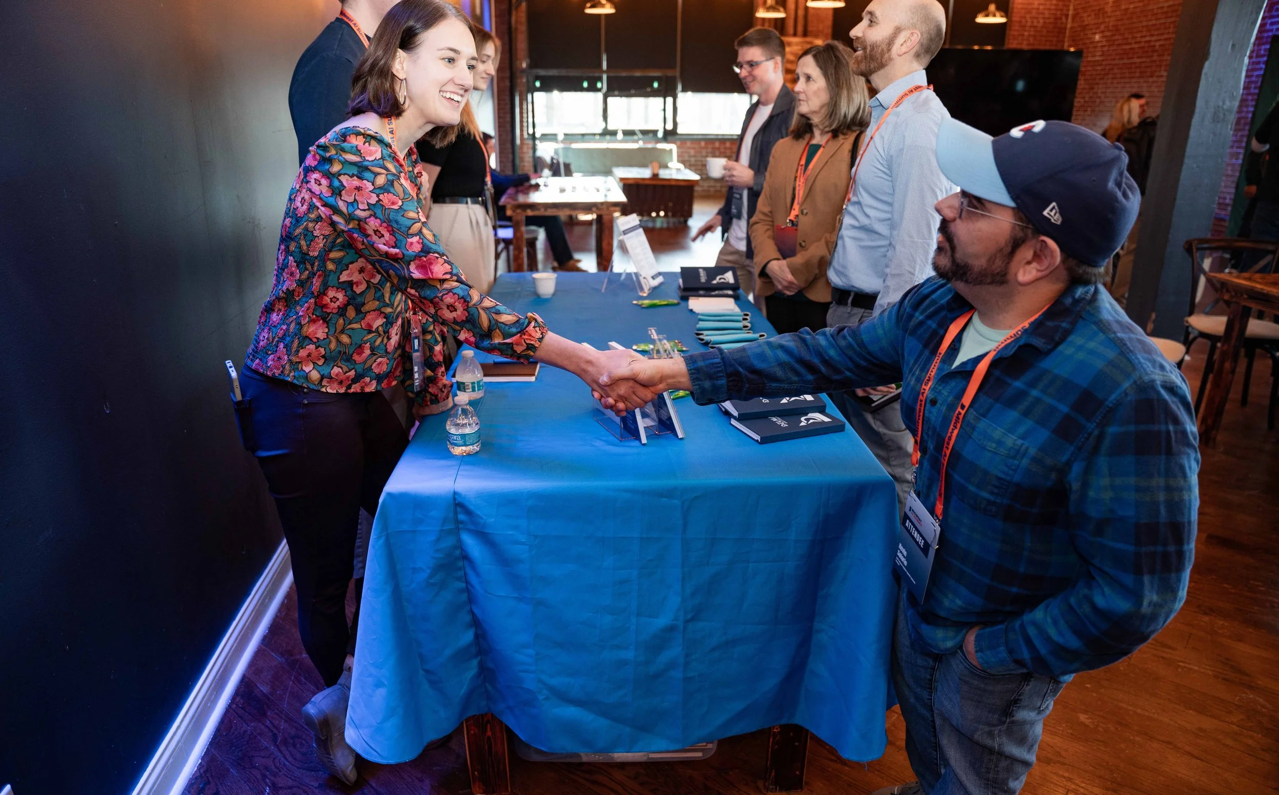 A woman in a floral jacket shakes hands with a man in a plaid shirt during a networking event at a registration table. Several other people are in line behind them, some holding coffee cups. The event is in a modern venue with brick walls and wooden 