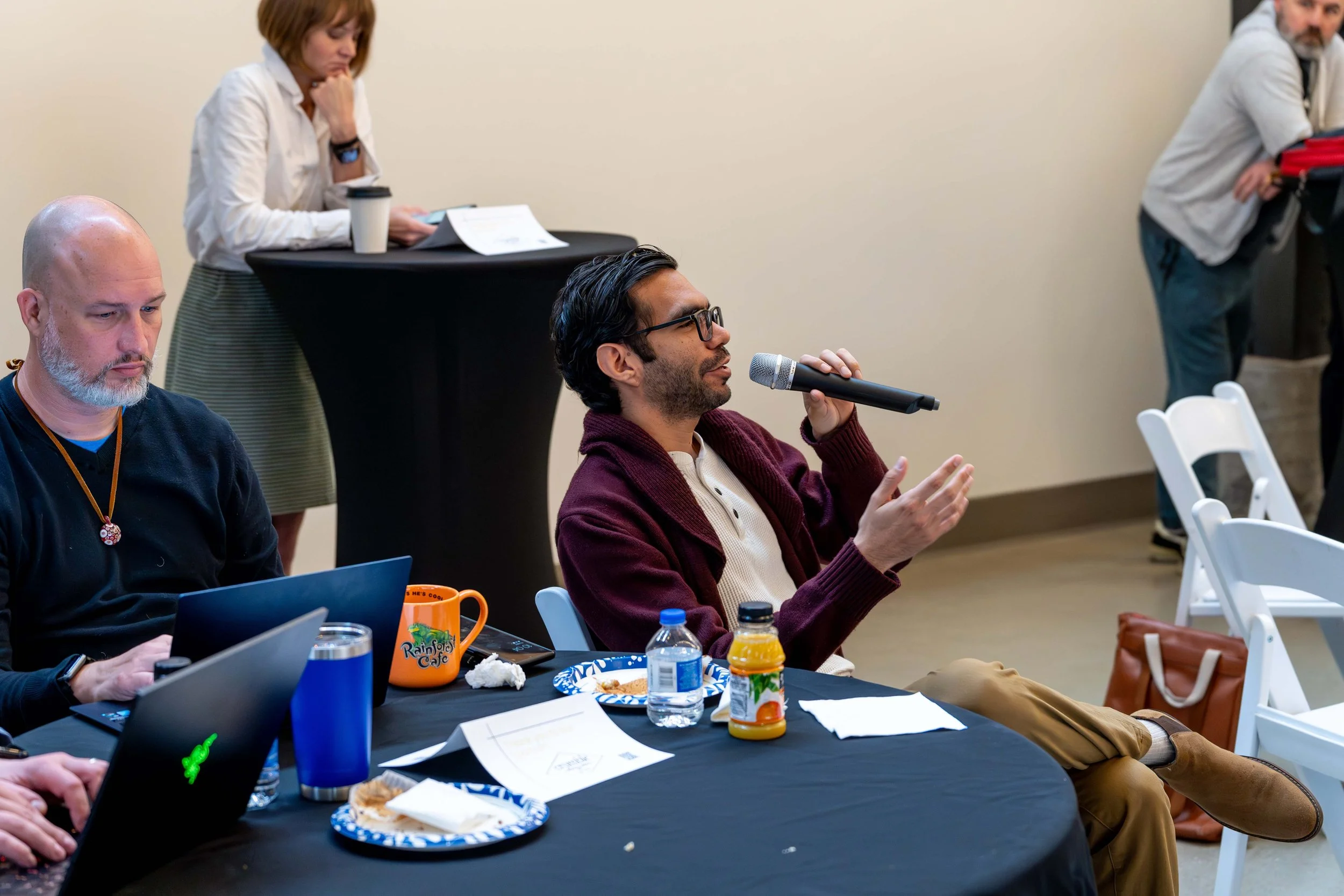 Man with dark hair and glasses wearing a maroon sweater, speaking into a microphone at a conference table with laptops, food, and drinks, while other attendees listen and work.