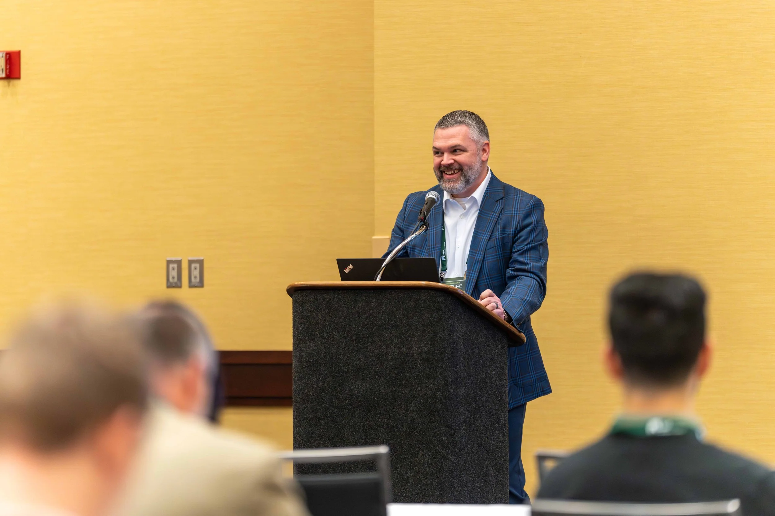 A man with gray hair and beard smiling while speaking at a podium during a conference or seminar, with an audience in front of him.
