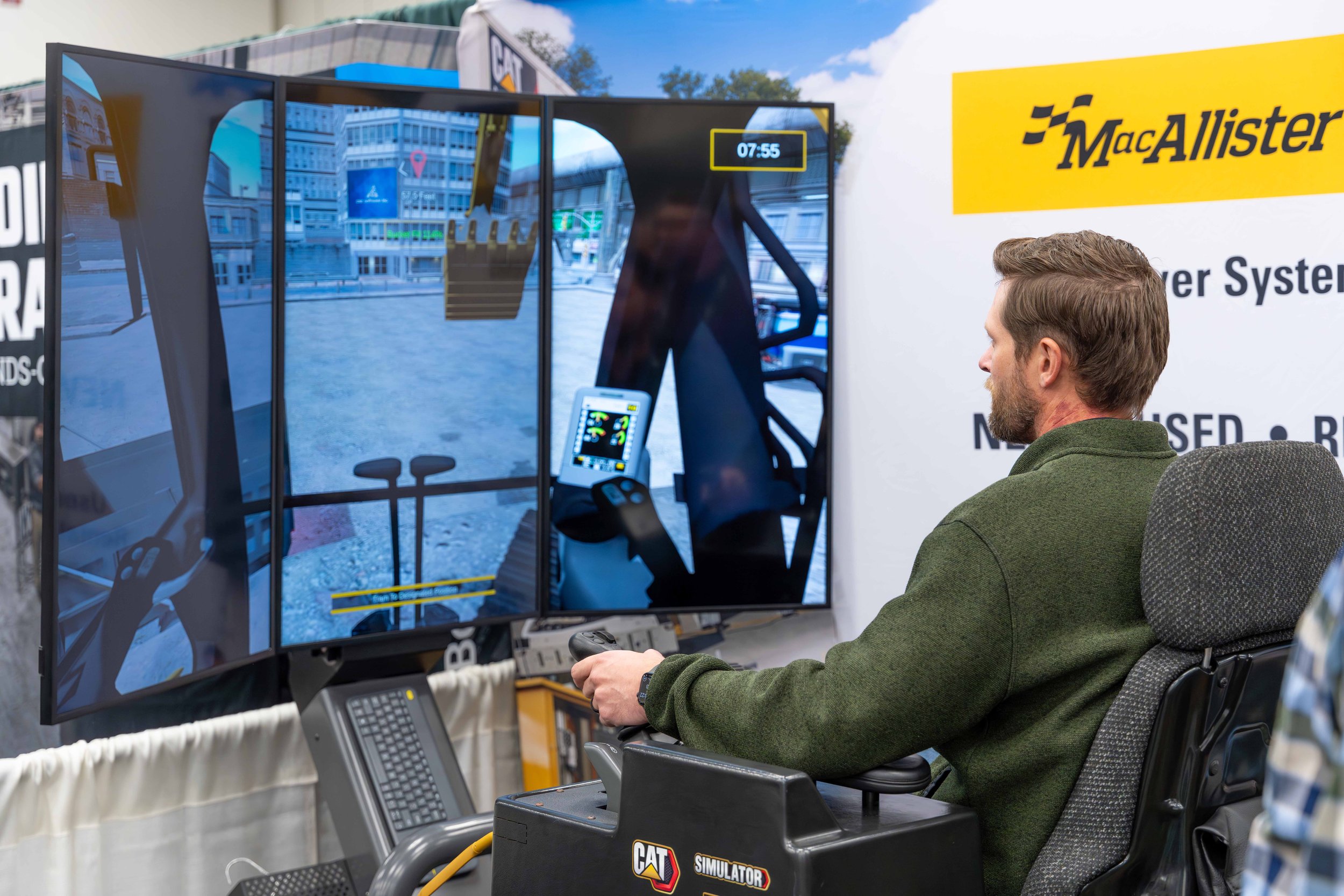 A man sitting in a simulator with large screens simulating a construction vehicle's driving view, surrounded by signage for MacAllister.