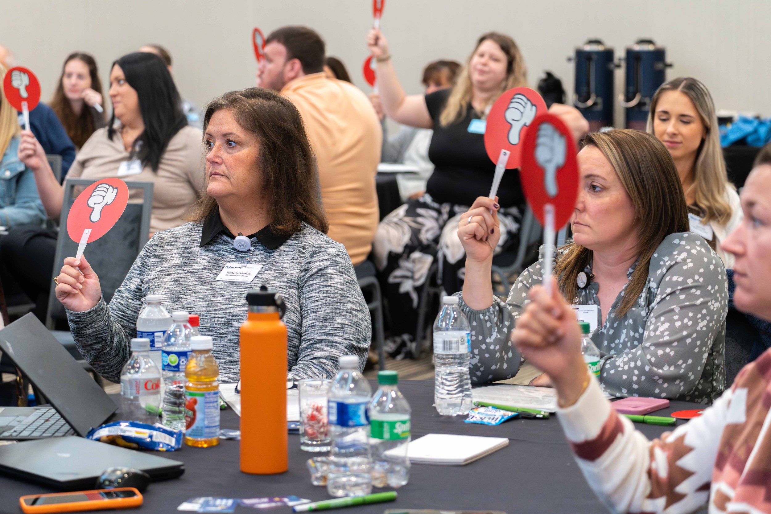 Conference room filled with people holding red signs with a thumbs-down symbol, seated at tables with water bottles, snacks, notebooks, and laptops.
