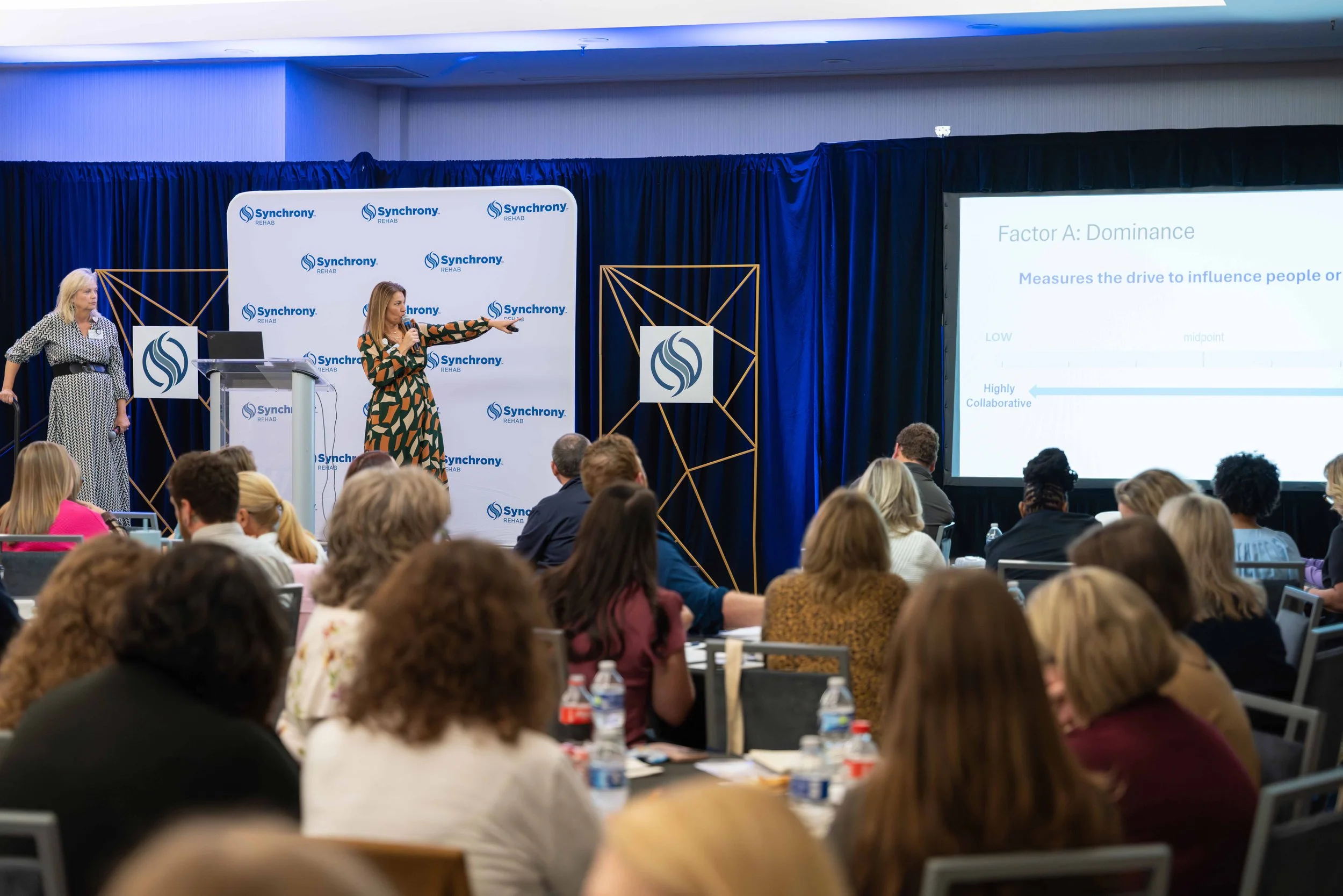 A woman in a patterned dress explains a presentation to an audience at a conference with a blue backdrop and a slide about dominance factors.