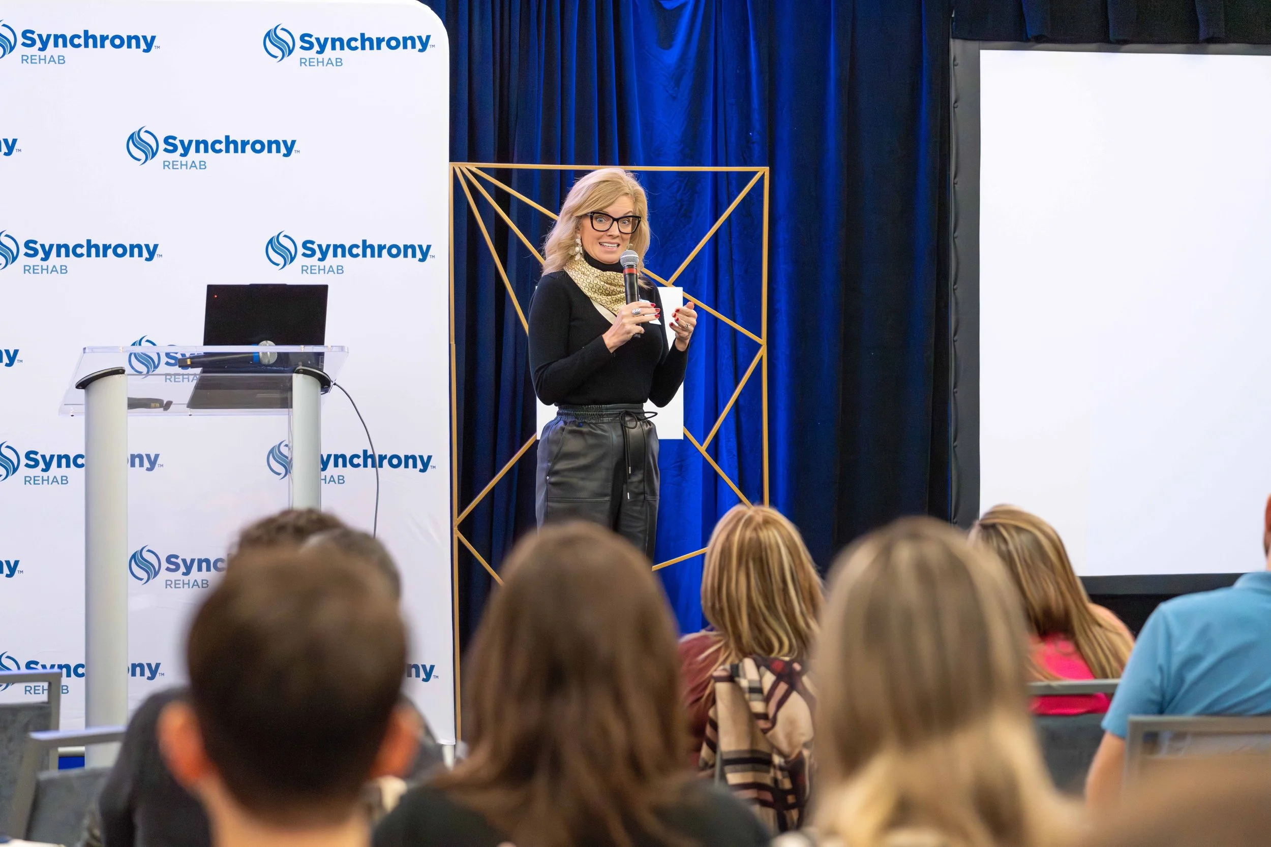 A woman with blonde hair, glasses, and a black outfit speaking at a conference with a microphone, standing in front of a blue curtain and a white screen with the 'Synchrony Rehab' logo, while an audience watches.