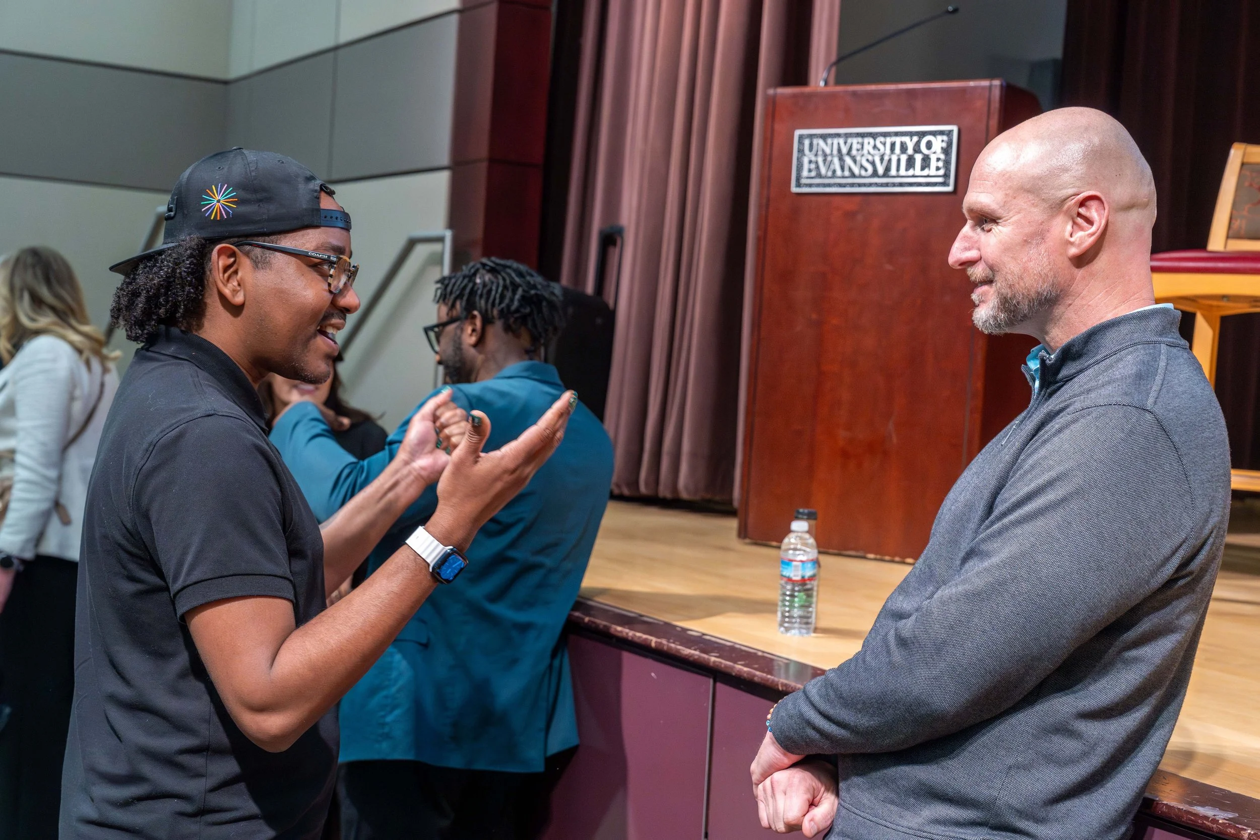 Two men having a conversation at an event, with a stage and a sign reading 'University of Evansville' in the background.