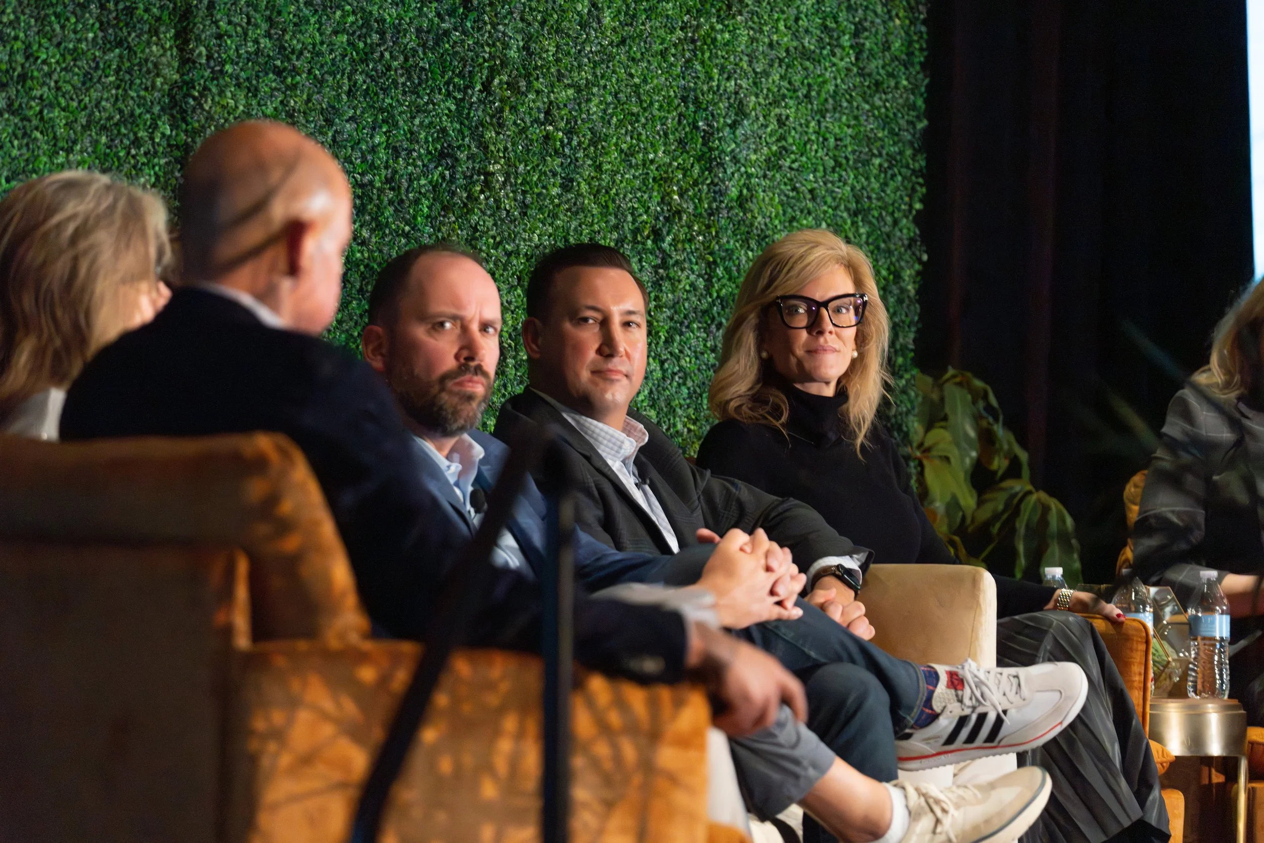 A panel of five individuals seated on a stage in front of a green leafy backdrop, participating in a formal discussion or conference.
