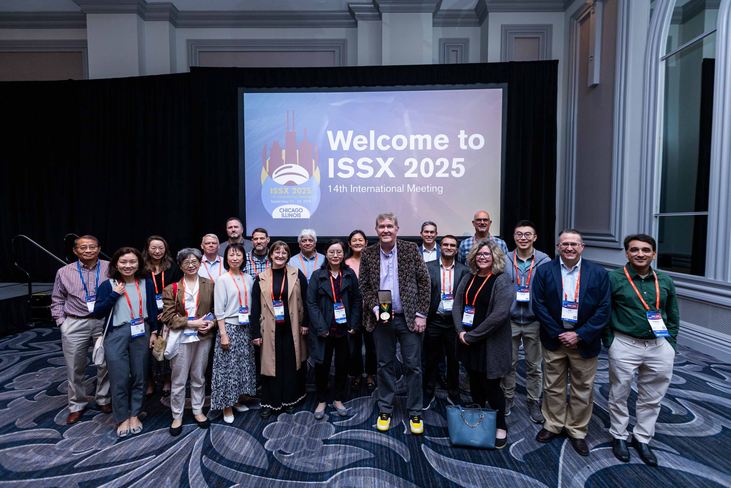 Group of diverse people gathered for a photo at the 14th International Meeting ISSX 2025 in Chicago, Illinois, standing in front of a large screen that displays event details.
