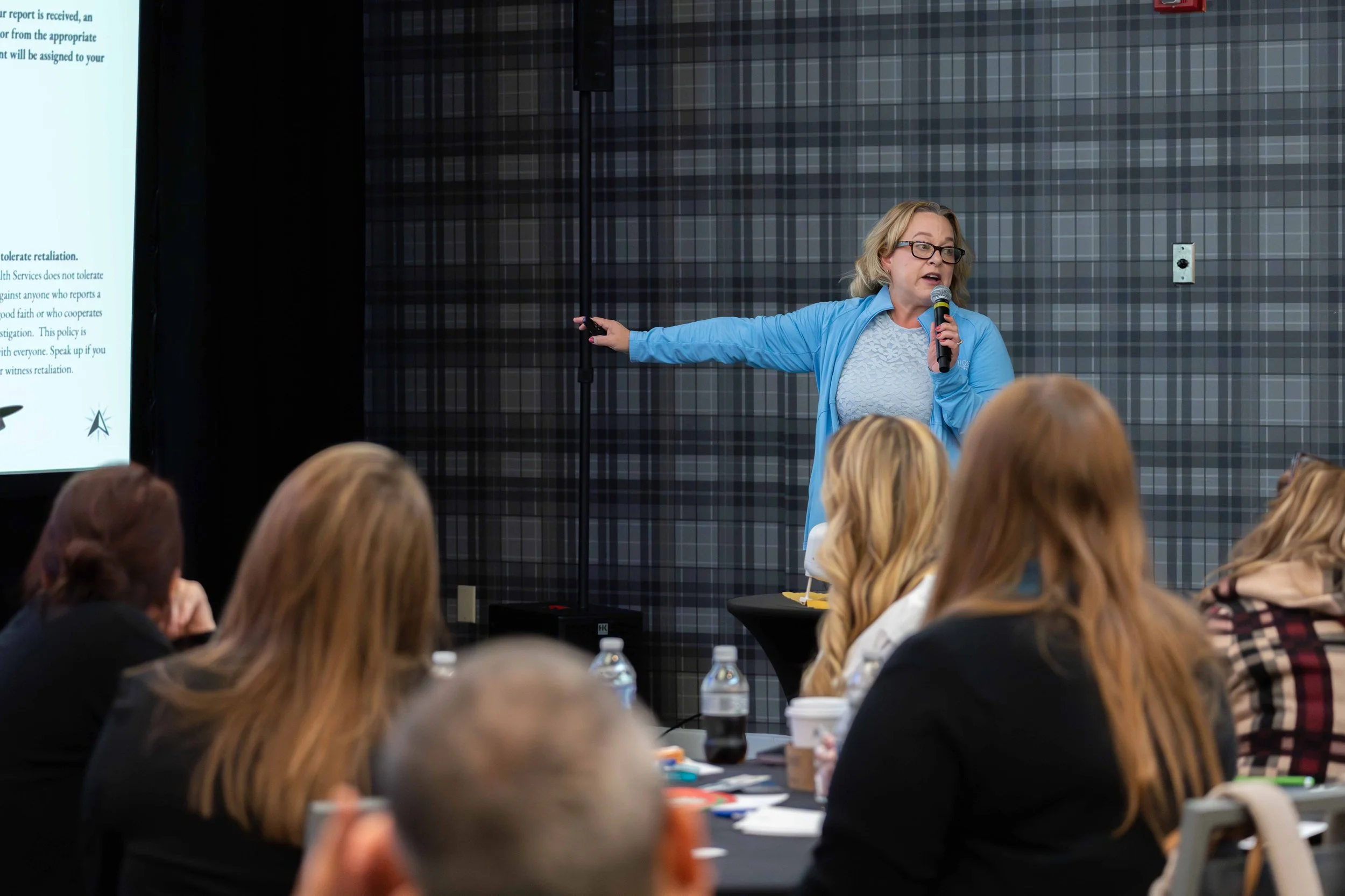 A woman in a blue jacket giving a presentation in front of an audience, holding a microphone in her right hand and pointing at a large screen with her left hand. The audience members are seated at tables, listening attentively.