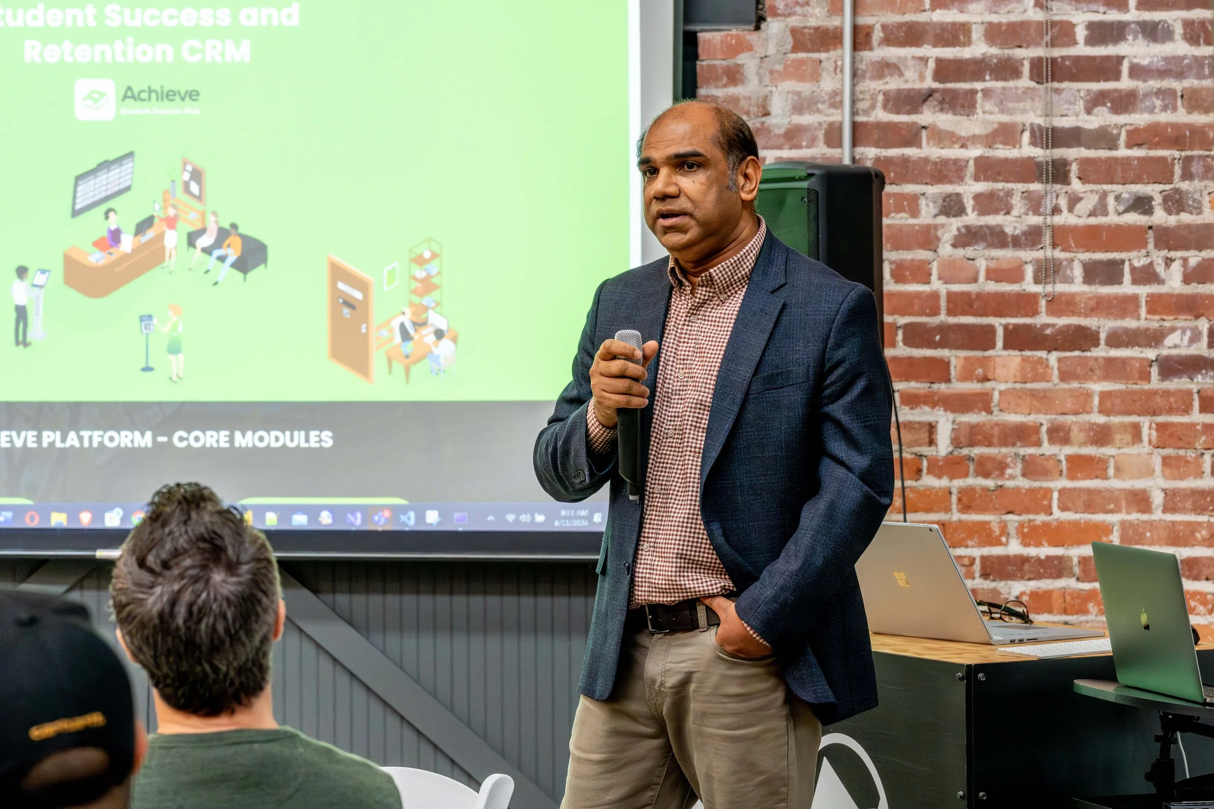 A man in a blazer and checked shirt holding a microphone, giving a presentation in a room with a brick wall, a projector screen displaying a slide about student success and retention CRM, and people seated listening.