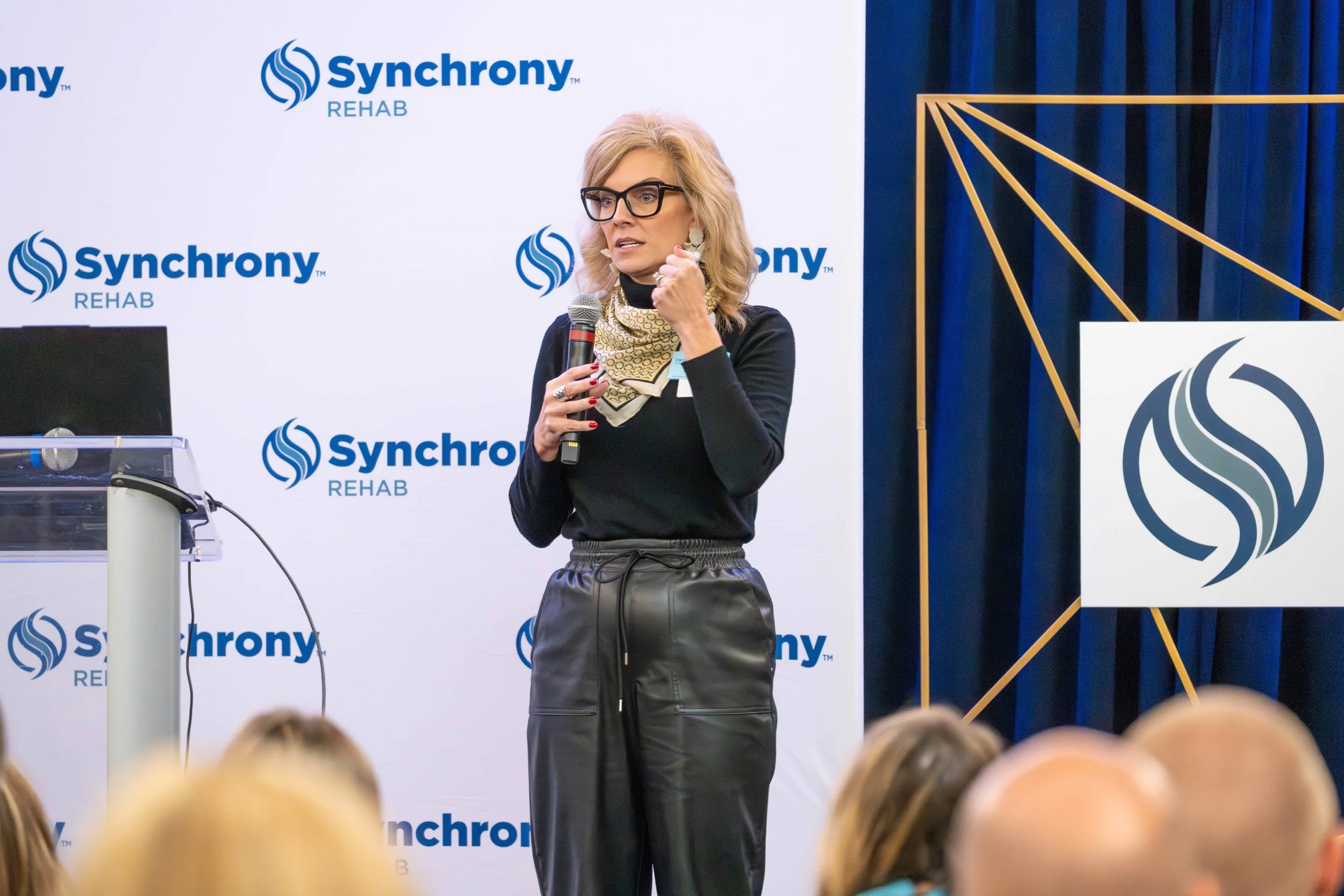 Woman speaking into a microphone at a conference with Synchrony Rehab branding in the background and a logo on the right side.