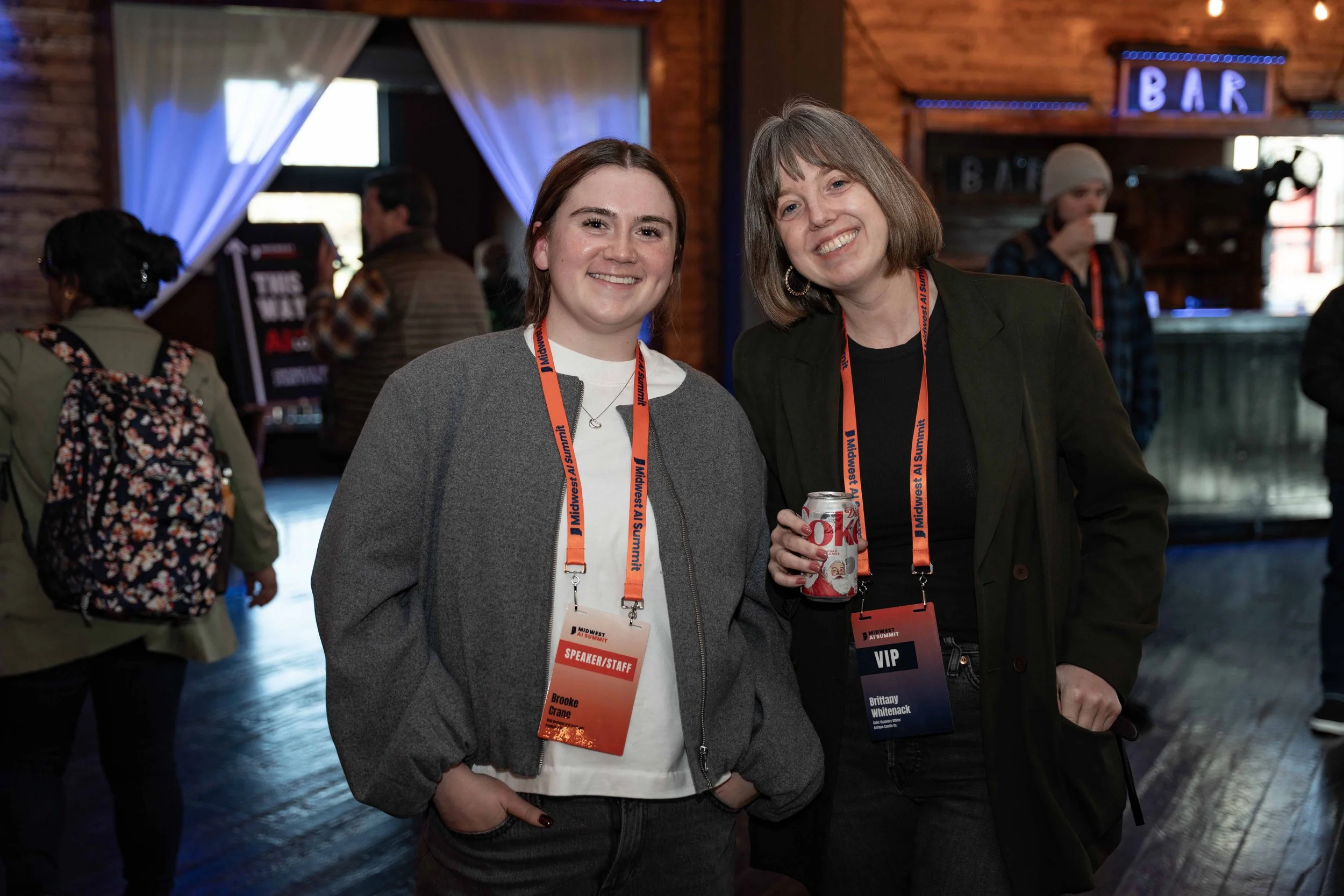 Two women smiling at a conference or event, both wearing lanyards with badges around their necks. One badge reads 'Speaker/Staff: Brooke Crane,' and the other's reads 'VIP: Brittany Whiteback.' The woman on the right is holding a can of Diet Coke. Th