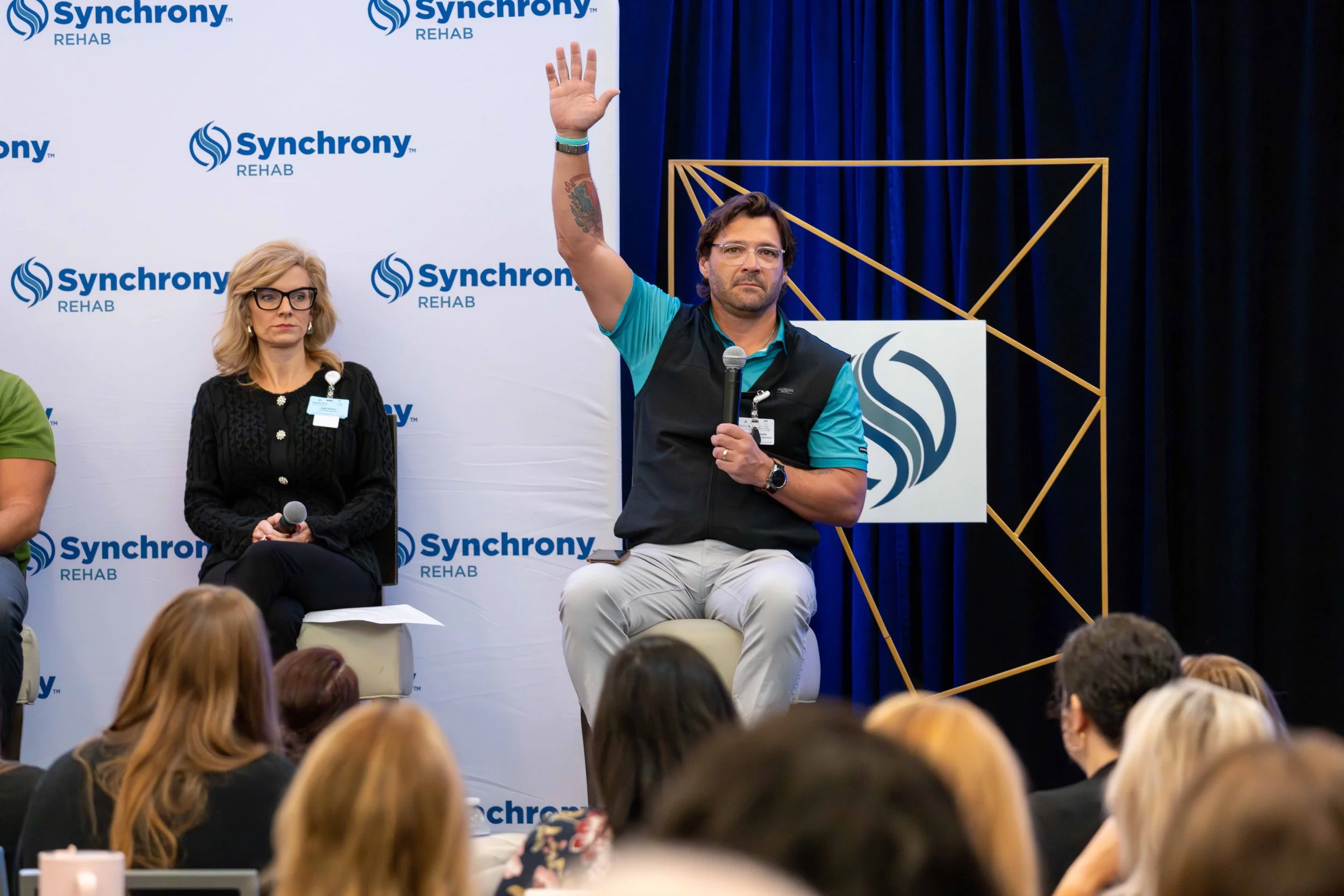 Man speaking at a conference, raising his hand, holding a microphone, seated on stage with a woman sitting beside him. Background has a banner with 'Synchrony Rehab' and a geometric gold frame.