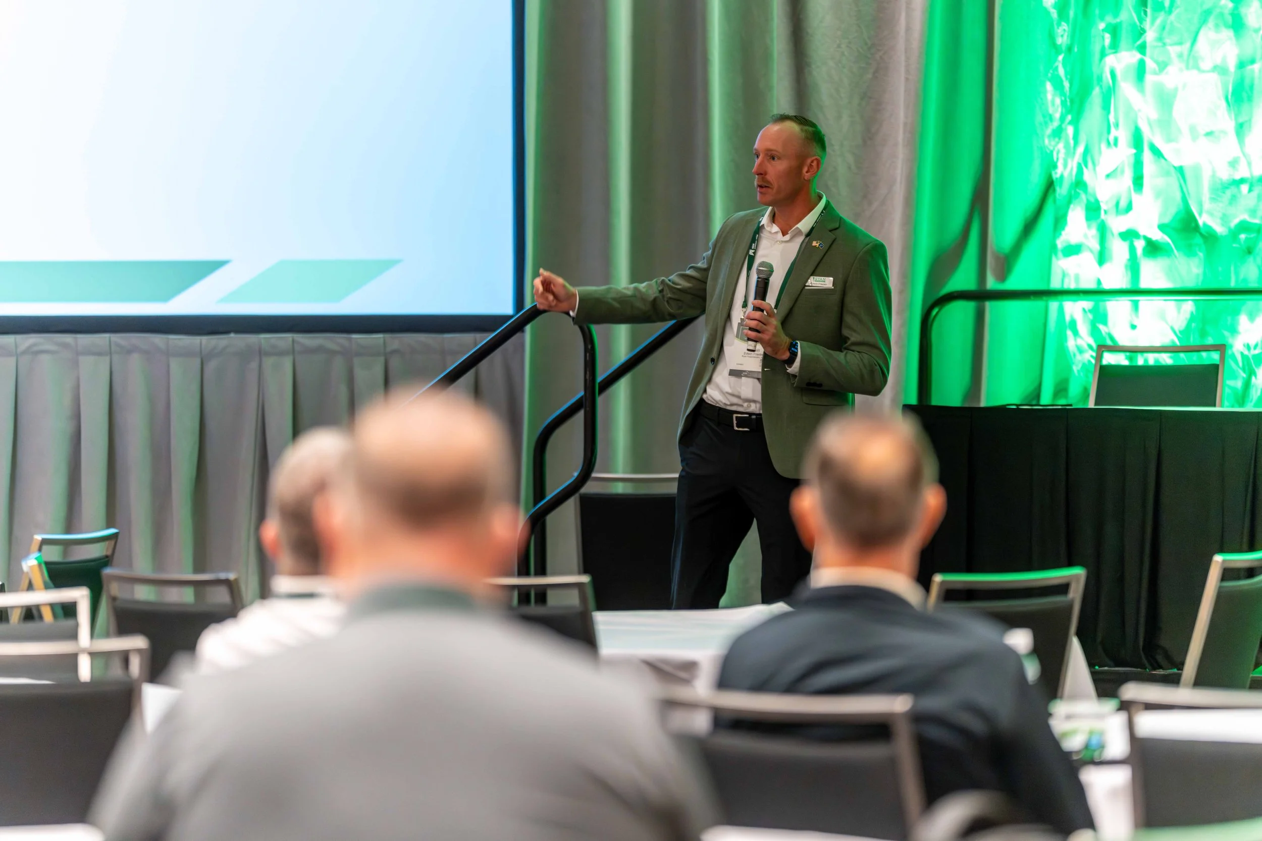 A man in a gray blazer and white shirt gives a presentation in front of an audience at a conference, holding a microphone.
