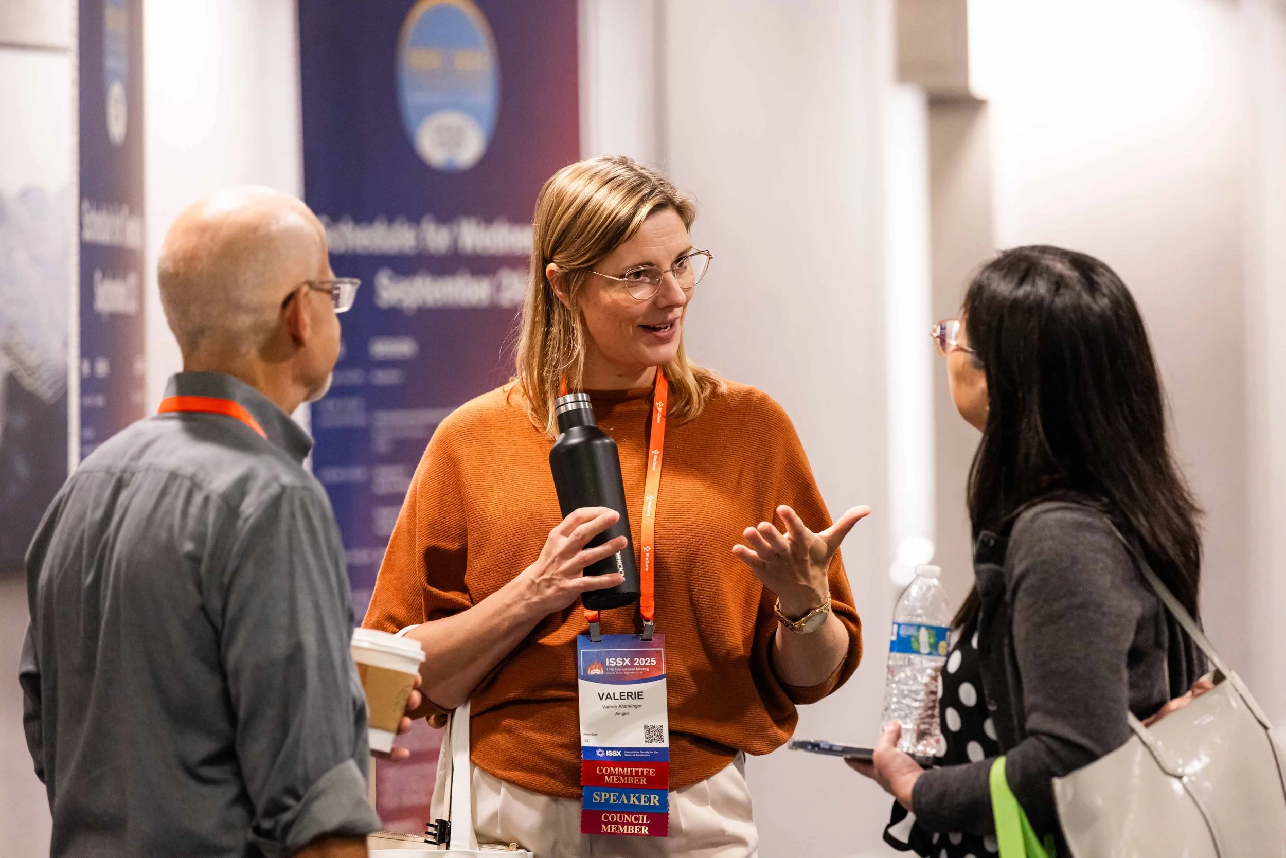 Three people engaged in conversation at a conference, two women and one man, with conference badges and a poster in the background.