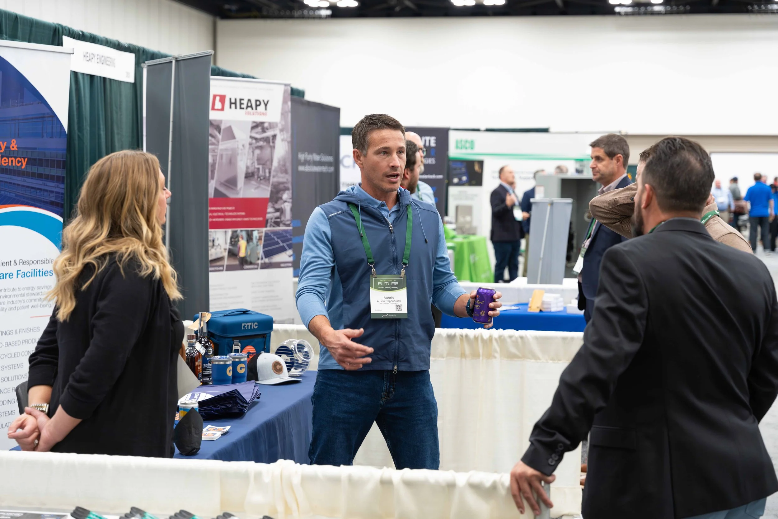 A man in a blue jacket and ID badge is speaking to a group of people at a trade show booth. The booth has promotional materials, and other attendees are visible in the background.