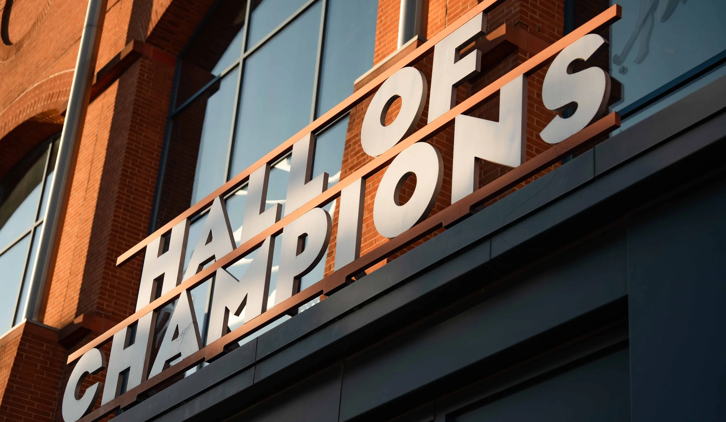 Sign on building reads 'CHALET OF CHAMPIONS' with a modern, reflecting glass exterior and red brick architecture in the background.