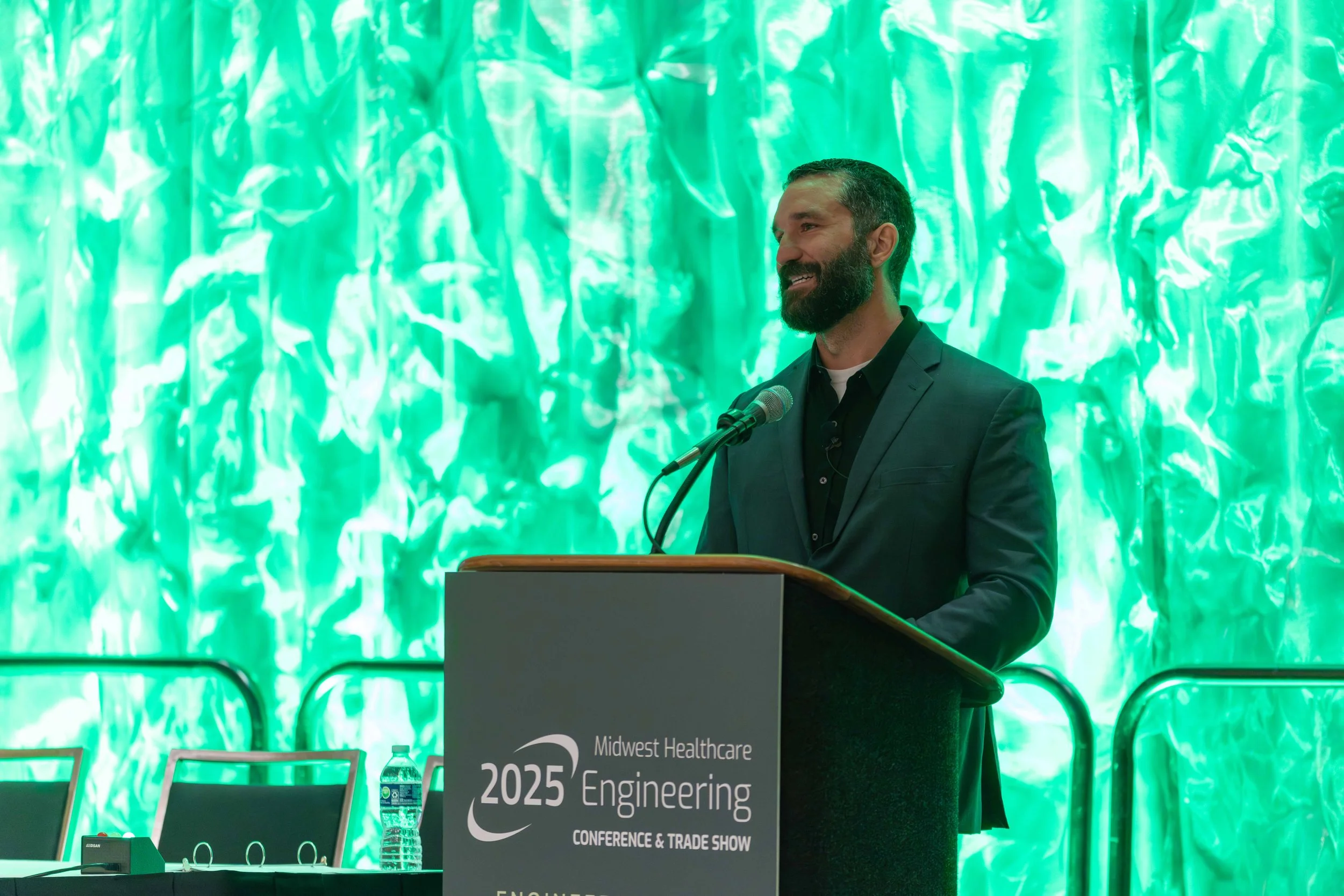 A man with a beard and short hair wearing a dark suit and black shirt standing behind a podium at a conference, smiling while speaking into a microphone. The background features a green textured backdrop.