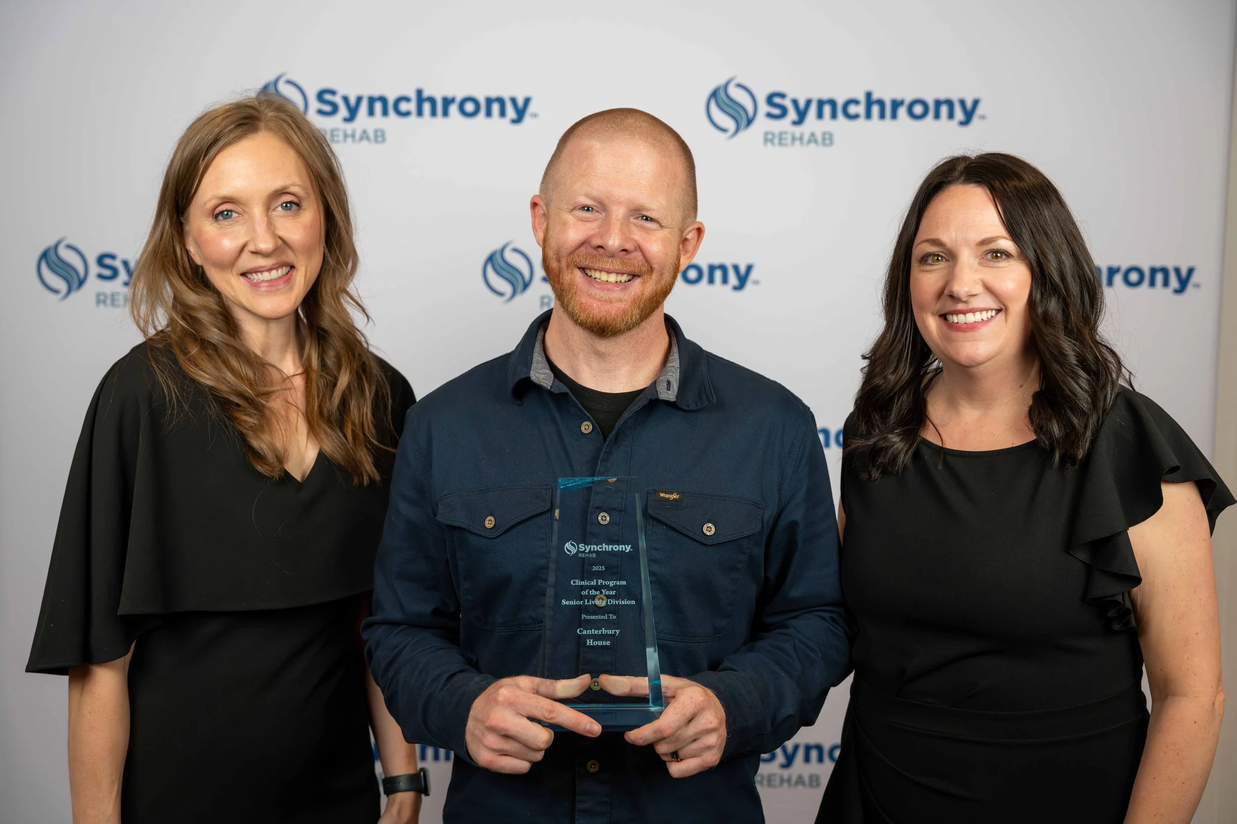 Three people standing together, one man in the middle holding an award, with a white backdrop displaying the Synchrony Rehab logo.